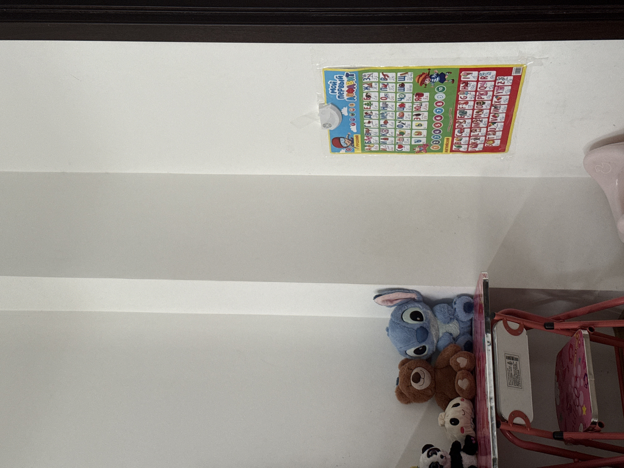 The image shows a corner of a room with a white wall and a dark brown door frame on the right. A small, pink step stool with a patterned seat is positioned beneath a shelf. On the shelf, there are several stuffed toys, including a panda, a round white character, a brown bear, and a blue Stitch character. To the right of the stuffed toys, a colorful Russian alphabet chart is taped to the wall. The chart displays letters, numbers, and illustrations. The lighting in the room appears to be natural and diffused, casting soft shadows. There is no visible indication of the specific location within Chișinău, Moldova.