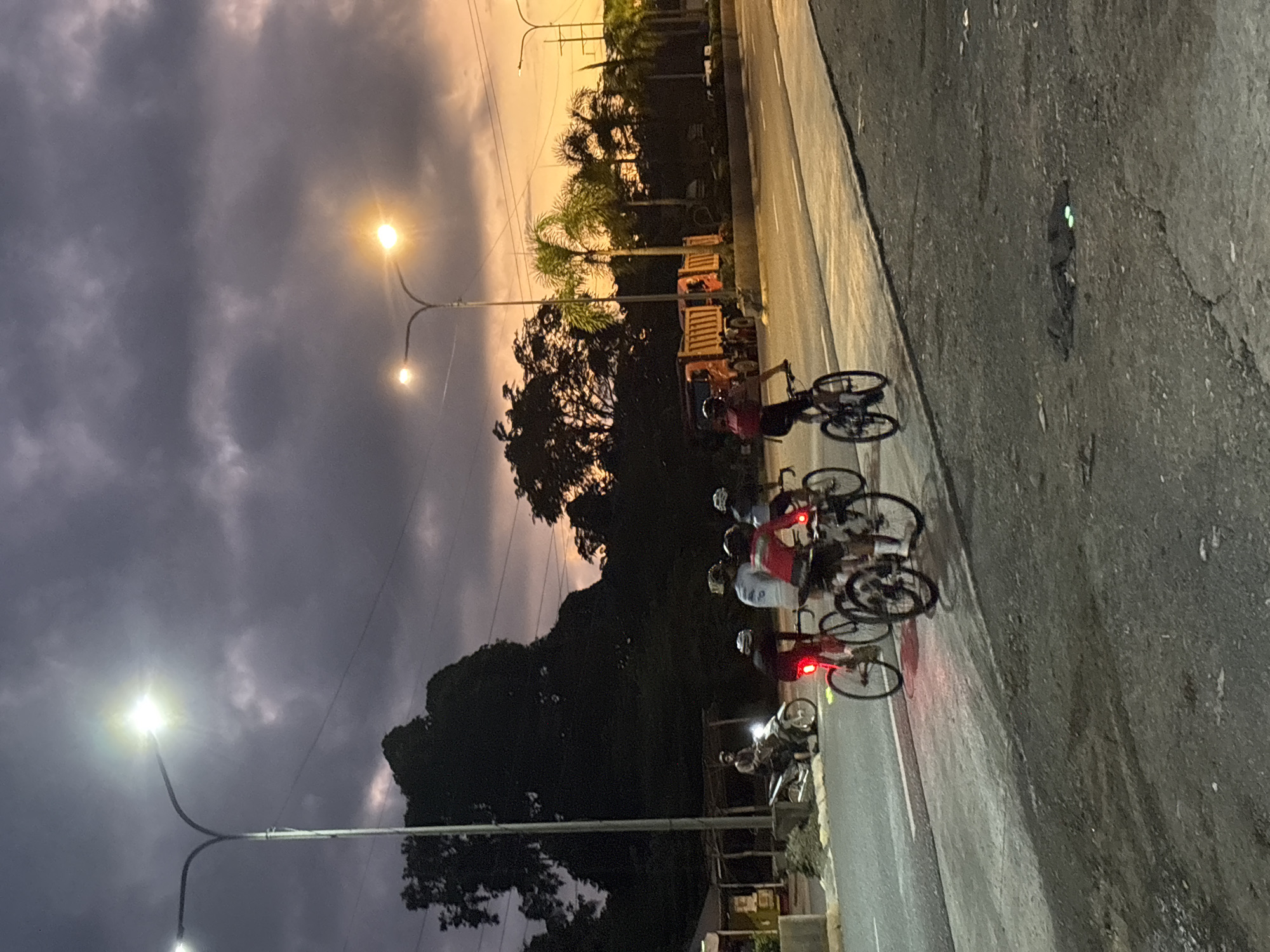 The image depicts a group of cyclists on a road during dusk. The sky is filled with dramatic clouds, transitioning from dark gray to shades of orange and yellow near the horizon. Several streetlights are illuminated, casting a warm glow. In the foreground, four cyclists are visible from the back, riding their bicycles. One cyclist in the lead wears a red shirt, followed by another in a light blue shirt, a third in a red shirt, and the fourth in a dark shirt. Their bicycles are equipped with red taillights. To the left, a person is seated on a motorbike, facing away from the camera. In the background, there are palm trees, a large dark tree, and two orange dump trucks parked on the side of the road. The road surface appears to be asphalt. The scene is set in the city of Carmen, Philippines.