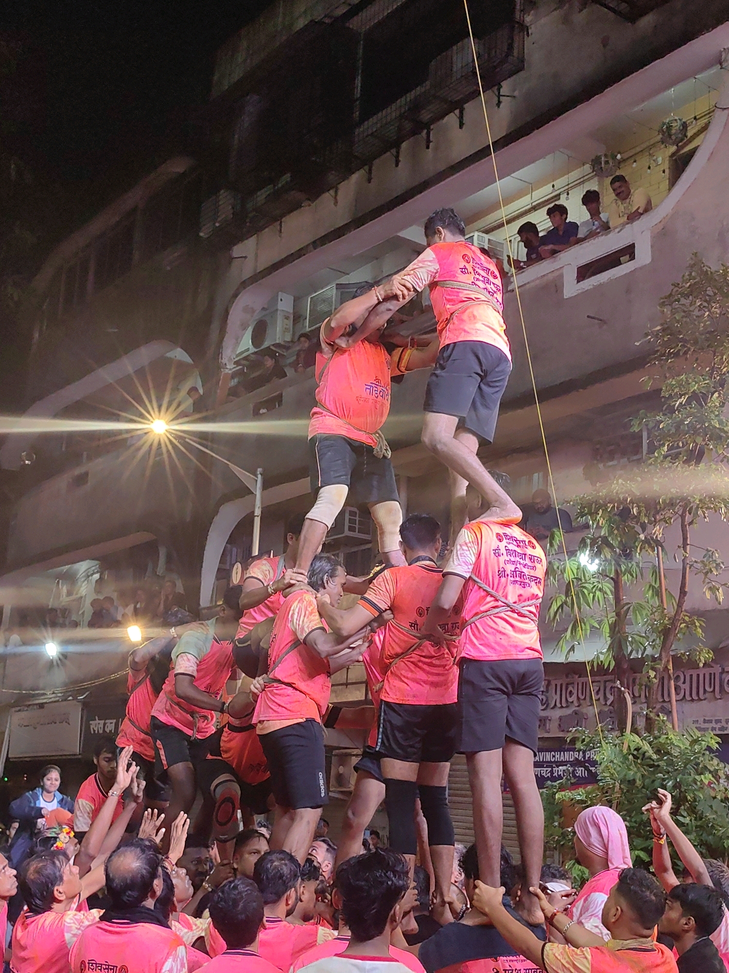 This image captures a human pyramid being formed by young men dressed in pink t-shirts and black shorts during a festival, likely Dahi Handi, in a densely populated urban setting in Mumbai, India. The pyramid is built in front of a multi-story building with balconies where spectators are visible, observing the event. The lighting suggests it is nighttime, with artificial lights creating a stark illumination on the participants. The environment appears to be a street or a public square, with trees and a building facade indicating a city environment. The participants in the pyramid are actively engaged in the physical exertion of climbing and supporting each other, with some individuals at the top reaching upwards. The crowd below is also engaged, with hands raised in support and encouragement. The overall scene depicts a lively and energetic cultural celebration.