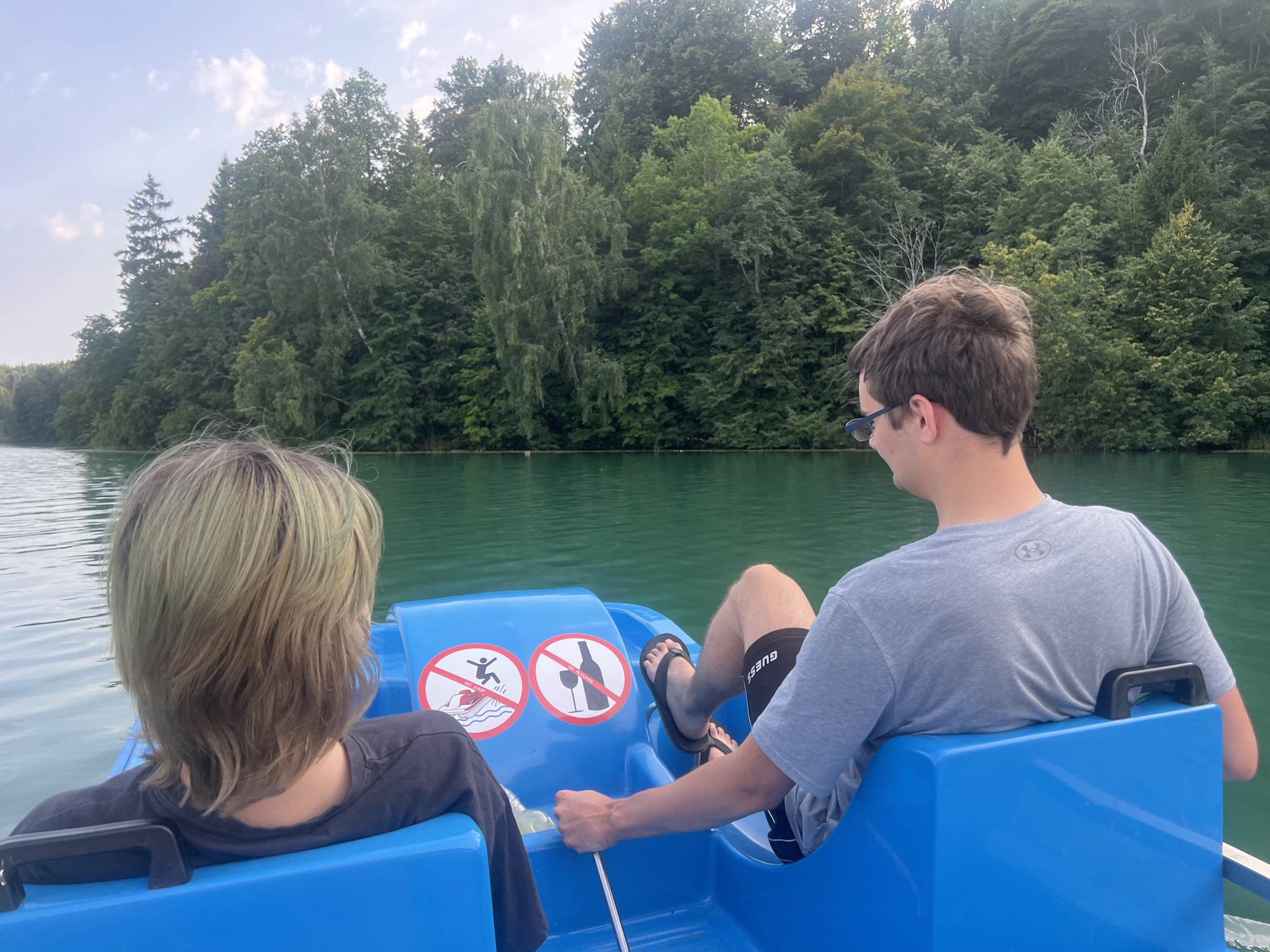 Two individuals are seated in a bright blue paddleboat on a tranquil body of water. The person in the foreground, facing away from the camera, has shoulder-length, light-colored hair and is wearing a dark t-shirt. Their hands are resting on the edge of the boat. To their right, a second person, also facing away, is wearing a grey t-shirt and black shorts. This person has short brown hair and is wearing sunglasses. They are actively operating the paddleboat, with their hands on the steering mechanism. The boat has two circular warning signs on its interior, one depicting a person jumping and the other showing a wine bottle and glass with a prohibition symbol. The water is a vibrant, clear green, reflecting the surrounding lush, dense forest that lines the opposite bank. The sky above is overcast with scattered white clouds. The overall scene suggests a leisurely outing on a lake or river, surrounded by nature. The image's origin is not specified and cannot be determined.