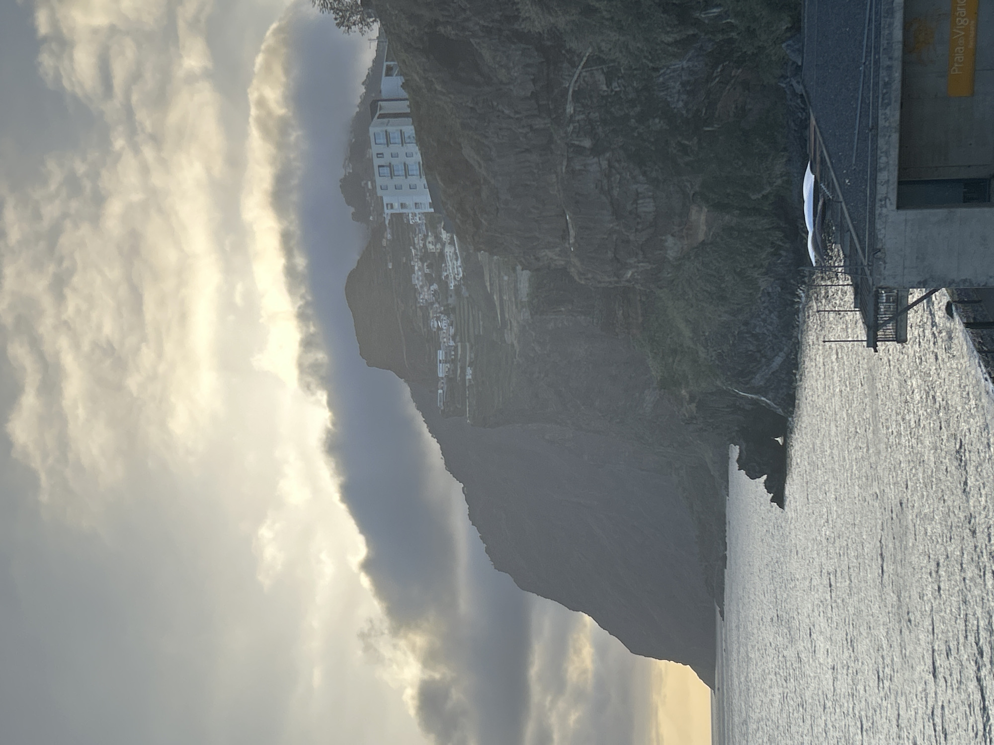 The photograph captures a coastal view in Câmara de Lobos, Portugal. A towering, rugged cliff dominates the scene, dotted with clusters of white buildings cascading down its steep slope. A dramatic sky filled with clouds, illuminated by the sun, is visible in the background. In the foreground, the ocean's surface is rippled with sunlight. A section of a man-made structure is visible on the right, bearing a sign with the inscription "Praia do Vigário". This suggests the location is near Vigário beach in Câmara de Lobos.