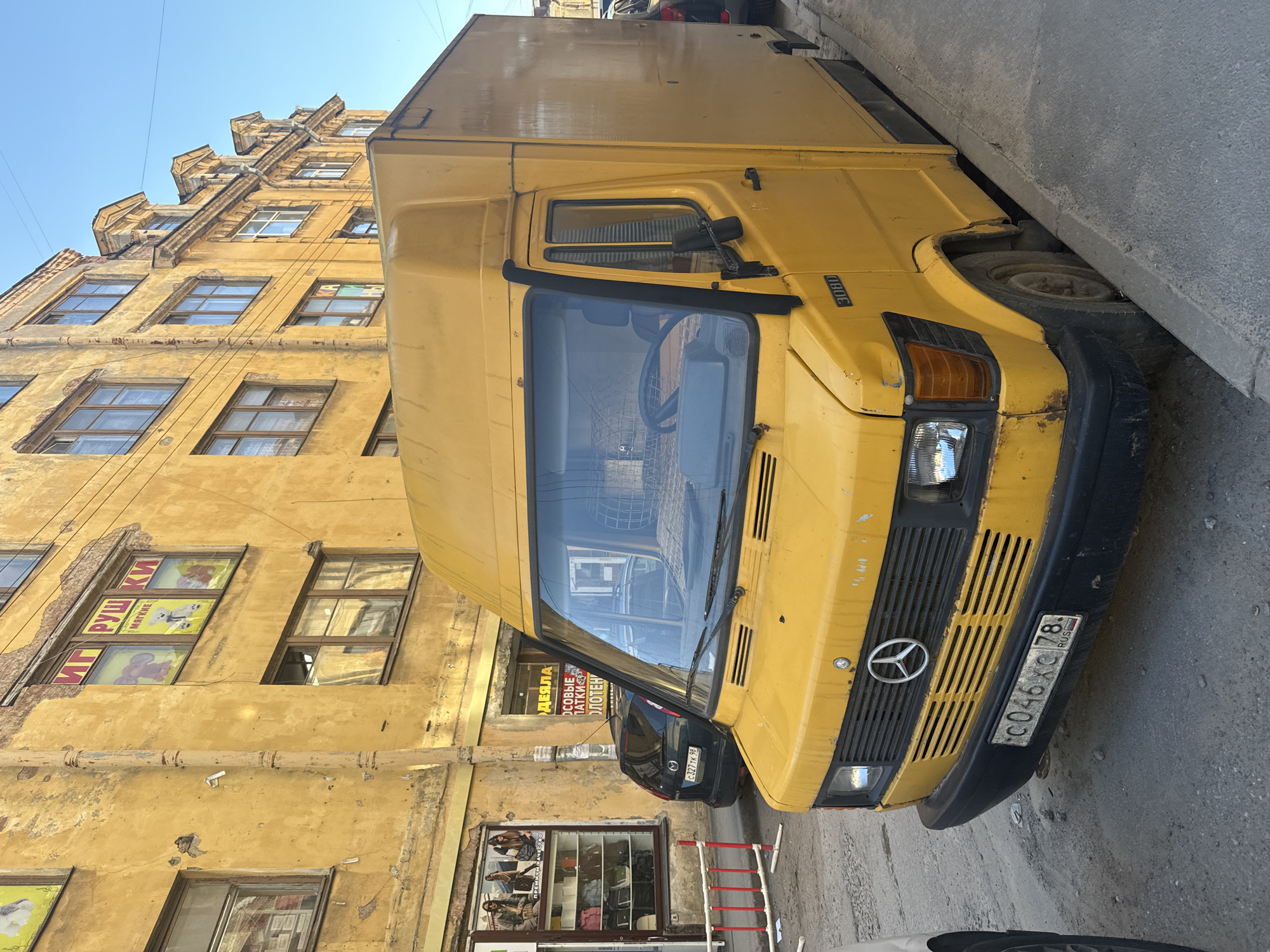 This is a photograph taken on a bright, sunny day in Saint Petersburg, Russia. The scene depicts a yellow Mercedes-Benz van parked on a narrow street, adjacent to a weathered, yellow building with multiple windows. The building shows signs of age, with peeling paint and some visible structural wear. Some of the windows display advertisements, including one for "МЯГКИЕ ПАТКИ" which translates to "soft ducklings" and likely indicates a toy store. Another window advertises "ОСОВЫЕ ПАТКИ ОЛОТЕН," which could be related to "soft ducklings" and "towels." The van has a license plate visible, "C046XC 78 RUS," with "78" indicating the Saint Petersburg region. The van itself has some wear and tear, with rust visible on the front bumper. A dark-colored car is partially visible behind the van. The composition is slightly tilted, giving a dynamic feel to the urban street scene. The overall impression is one of everyday urban life in an older part of the city, bathed in clear daylight.