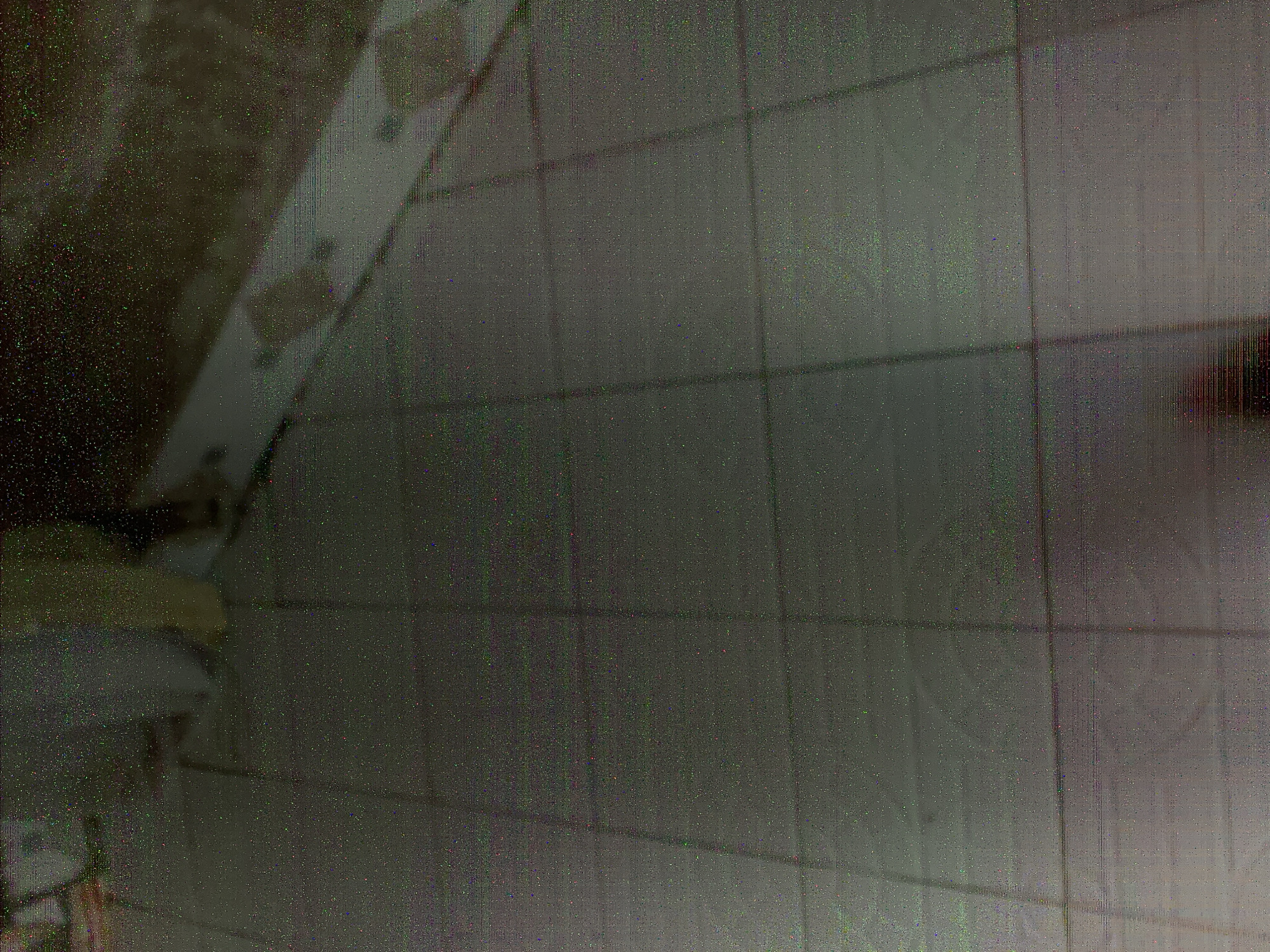The image depicts a tiled floor. The tiles are square and laid in a grid pattern. The grout lines between the tiles are visible. The lighting in the scene is dim, and there is a significant amount of digital noise and color distortion throughout the image. In the upper left corner, there is a blurred area with what appears to be part of a person or object. The rest of the image is dominated by the texture and pattern of the floor tiles, which are rendered in varying shades of gray and beige. The overall impression is one of low-resolution and poor image quality, making it difficult to discern specific details beyond the tiled surface.
