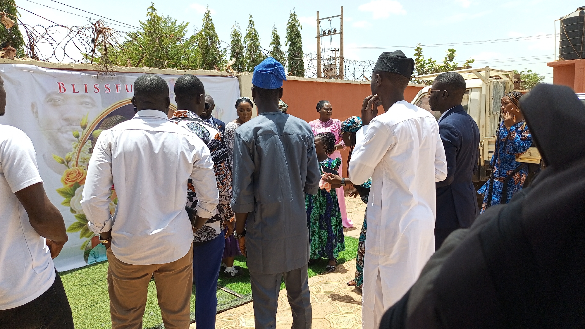 Kakau, Nigeria: A group of approximately 15 people are gathered outdoors near a banner with the word "Blissful" printed on it. The banner also features a photo and is decorated with roses. The people are dressed in a variety of clothing styles and colors. The environment includes trees, a building, and what appears to be a barbed wire fence. One person is holding a phone.