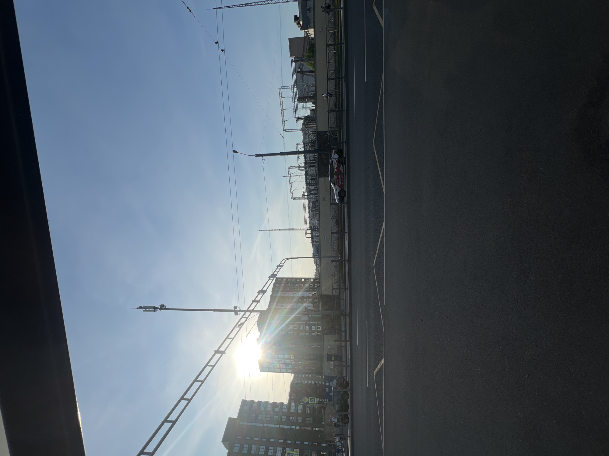 The image depicts a street scene in Saint Petersburg, Russia, likely during daylight hours, given the strong sunlight. In the foreground, a dark asphalt road with white lane markings is visible, suggesting a multi-lane thoroughfare. Beyond the road, a pedestrian walkway or sidewalk is bordered by a metal fence.
On the other side of the fence, several elements contribute to an urban industrial landscape. There are tall, modern residential buildings with multiple windows, one of which is prominently illuminated by the sun, creating a lens flare effect. Adjacent to these buildings is an electrical substation, characterized by numerous metal structures, wires, and poles. A cell tower is also visible, a common feature in urban environments.
A few vehicles are present on the road. One car, a light-colored sedan with red and white markings, is positioned near the substation. Another dark-colored car is seen closer to the residential buildings. Several pedestrians are visible in the distance, either walking on the sidewalk or near the buildings. The sky is clear and blue with a few faint clouds, and the sun is high, indicating it's likely daytime. The overall impression is of a bustling urban environment with a mix of residential, commercial, and industrial infrastructure.