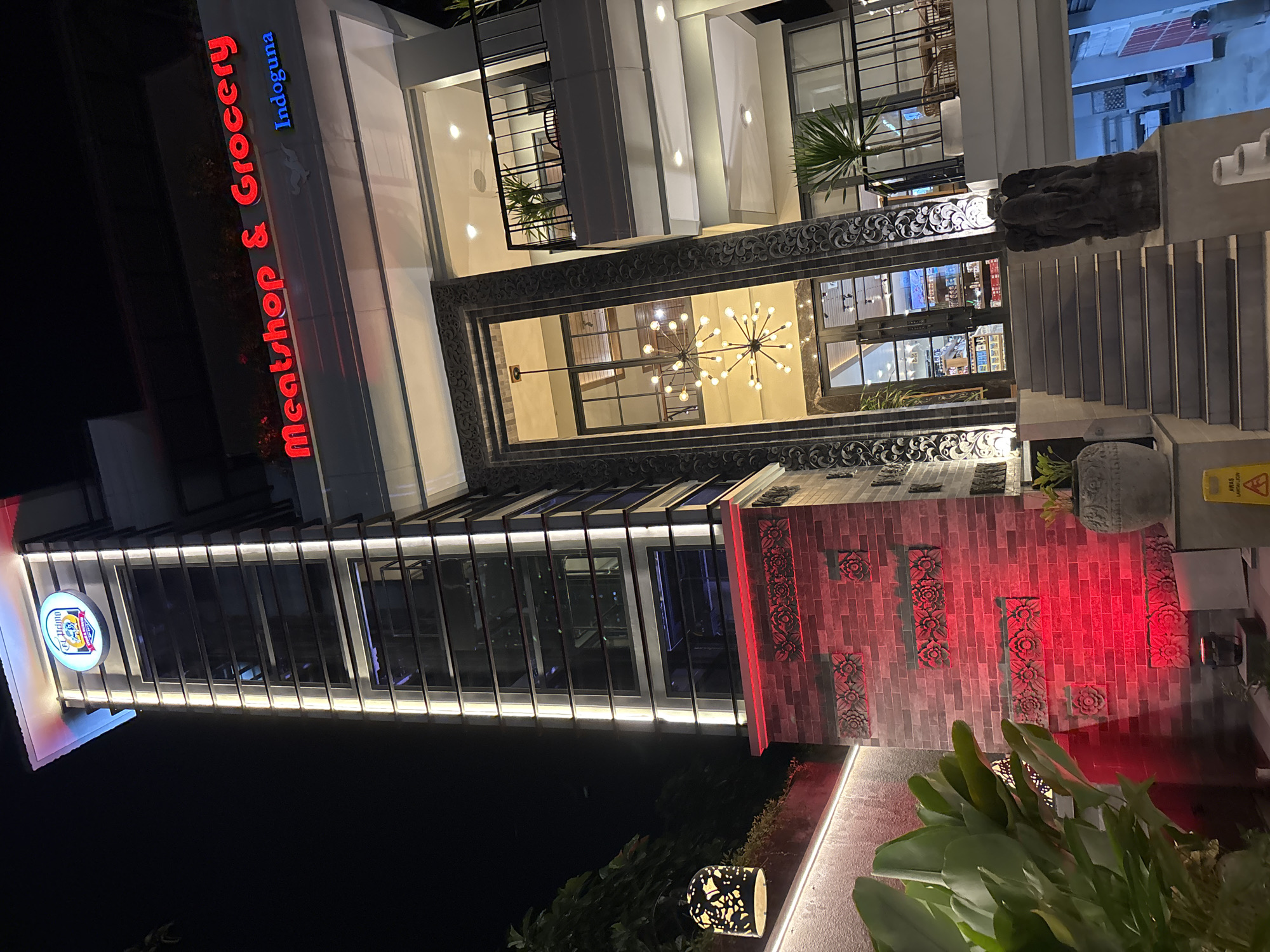 The image depicts the exterior of "Meatshop & Grocery Indoguna," a building located in Kuta Selatan, Indonesia, at night. The building features a modern design with multiple levels, large windows, and decorative elements, including brickwork and ornamental borders. A brightly lit sign displays the store's name. A staircase leads up to the entrance, and greenery is present near the base of the building. A decorative lamp provides additional illumination.