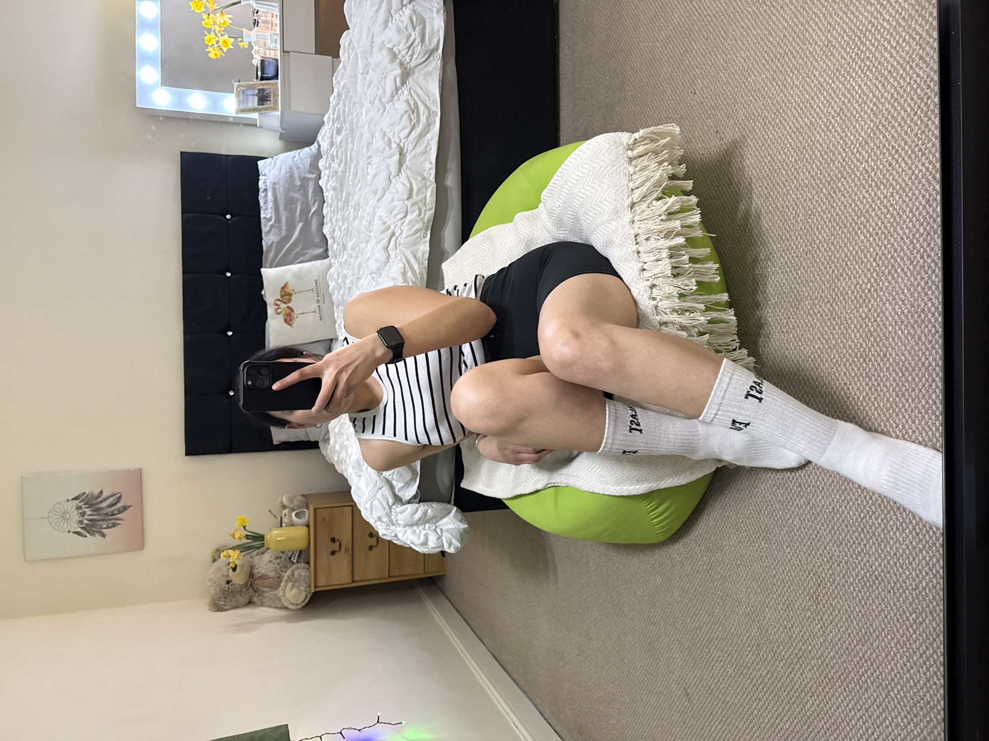 A person is photographed in a bedroom. The person is sitting cross-legged on a green beanbag chair, wearing black shorts, a striped black and white tank top, and white socks with black lettering. The person is holding a black smartphone and taking a picture. A bed is in the background with a black headboard, white comforter, and several pillows. To the left of the bed is a wooden nightstand with a teddy bear and flowers on top. On the wall above the nightstand is a framed picture. A makeup vanity with a light-up mirror is also visible. The room has beige carpet. It is impossible to identify the location as Great Yarmouth, United Kingdom, with the provided information.