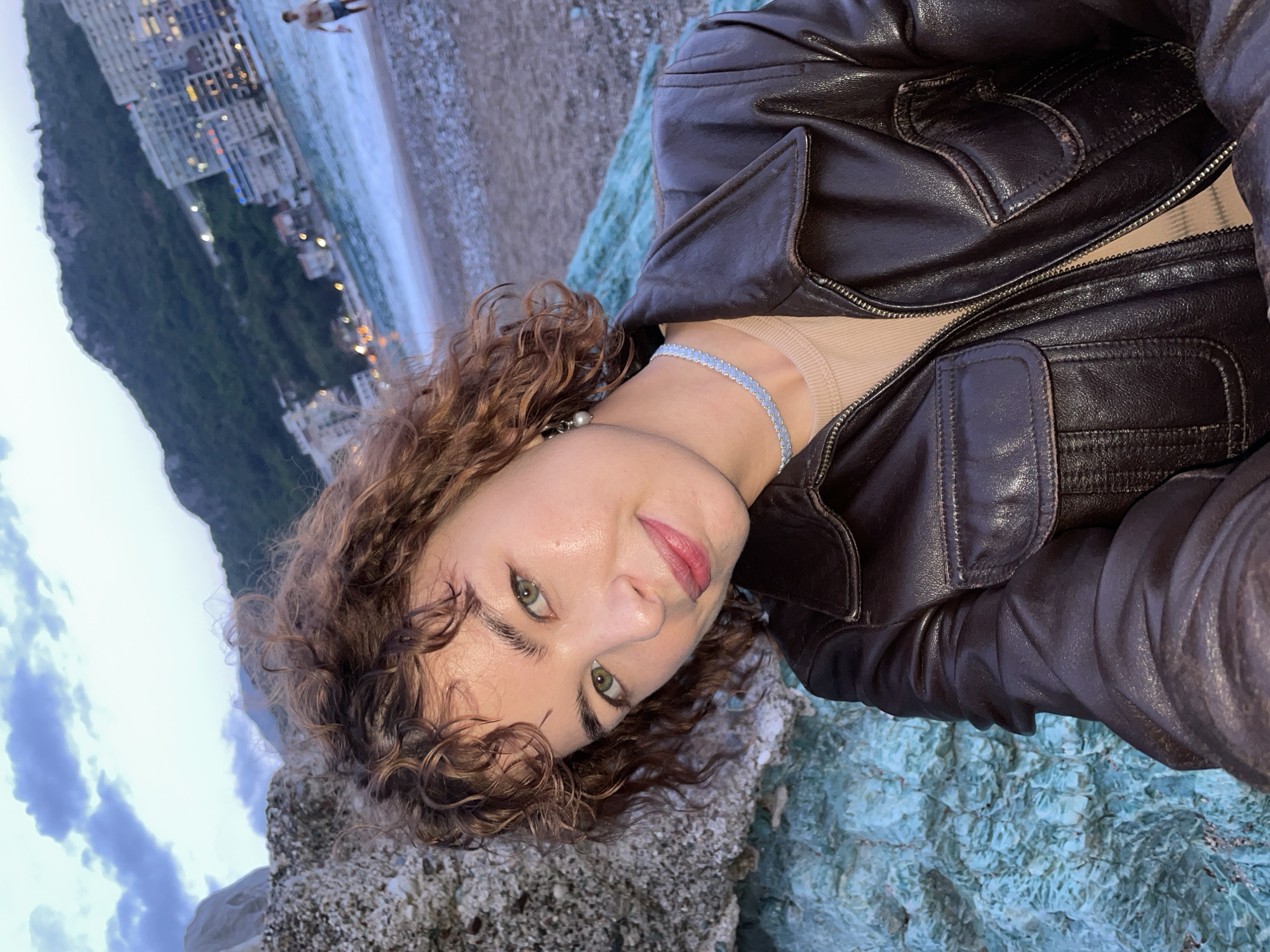 A selfie captures a young woman with curly auburn hair, light skin, and green eyes. She is wearing a brown leather jacket and a light blue choker necklace. The background reveals a beach scene. In the distance, buildings are visible, illuminated by lights, along with a mountain. There is also a person on the beach walking into the water. The woman appears to be positioned on rocky, turquoise-colored surface. Based on the buildings and coastline, the location is likely Rafailovići, Montenegro.