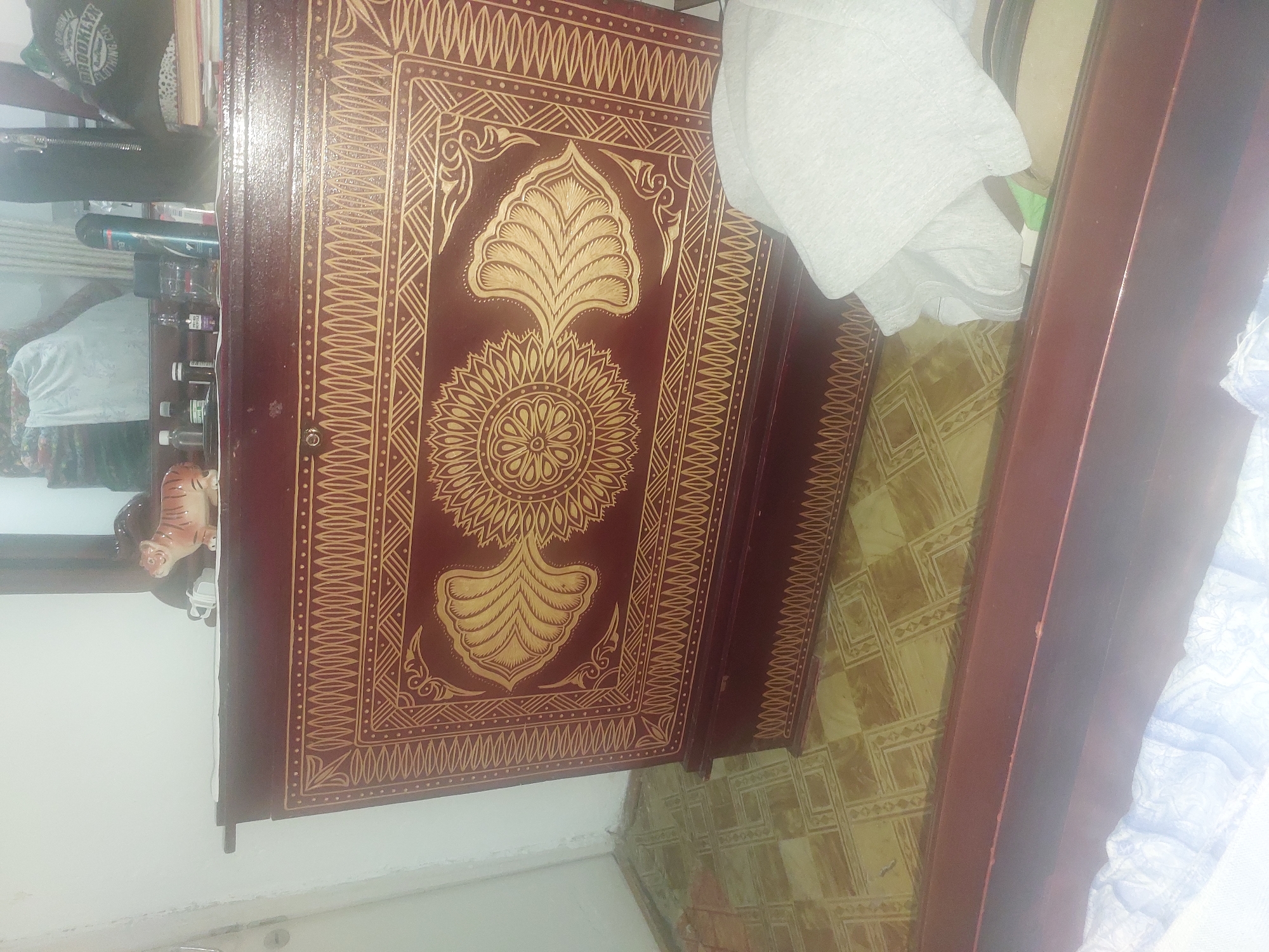 A dark red wooden chest with intricate gold geometric patterns and a floral design is positioned on a tiled floor. A tiger figurine, white towels, and other various objects are on top of the chest, suggesting an indoor setting. The scene is dimly lit, creating shadows and reflections on the surfaces. The location is identified as Ması, Kyrgyzstan.