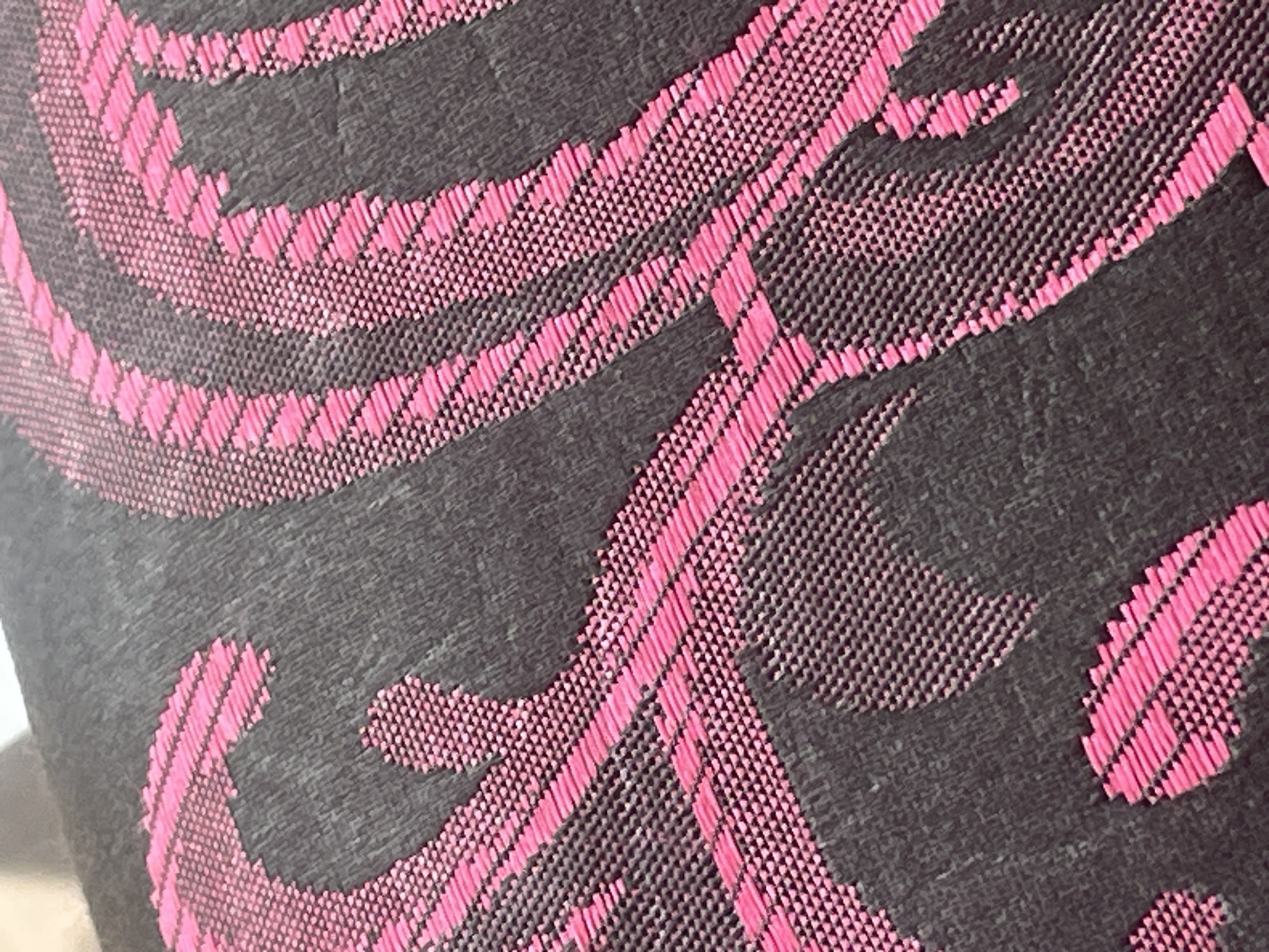 The image shows a close-up of a dark gray fabric with a raised, woven pattern in bright pink. The pattern consists of flowing, curvilinear lines that resemble abstract floral or paisley motifs. The texture of the fabric is visible, with small, uniform woven threads forming both the background and the raised pattern. The lighting highlights the texture and sheen of the pink threads. There is no indication of a specific location in Jalingo, Nigeria, or any people, actions, or interactions. The image is a detail shot of a textile.