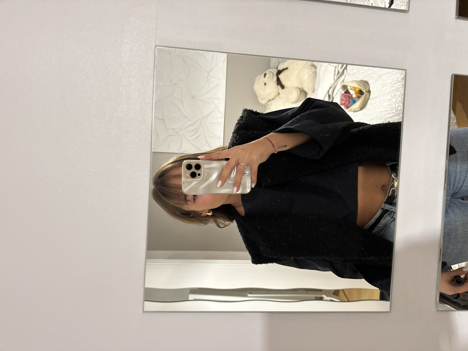 A reflection in a square mirror shows a woman taking a selfie. She is wearing a black top and a furry jacket. The phone has a pearly white case. Parts of her jeans are visible. Behind her reflection is a teddy bear on a white bed with other objects. A white wall is to the left of the mirror. Another partially visible mirror is on the right.