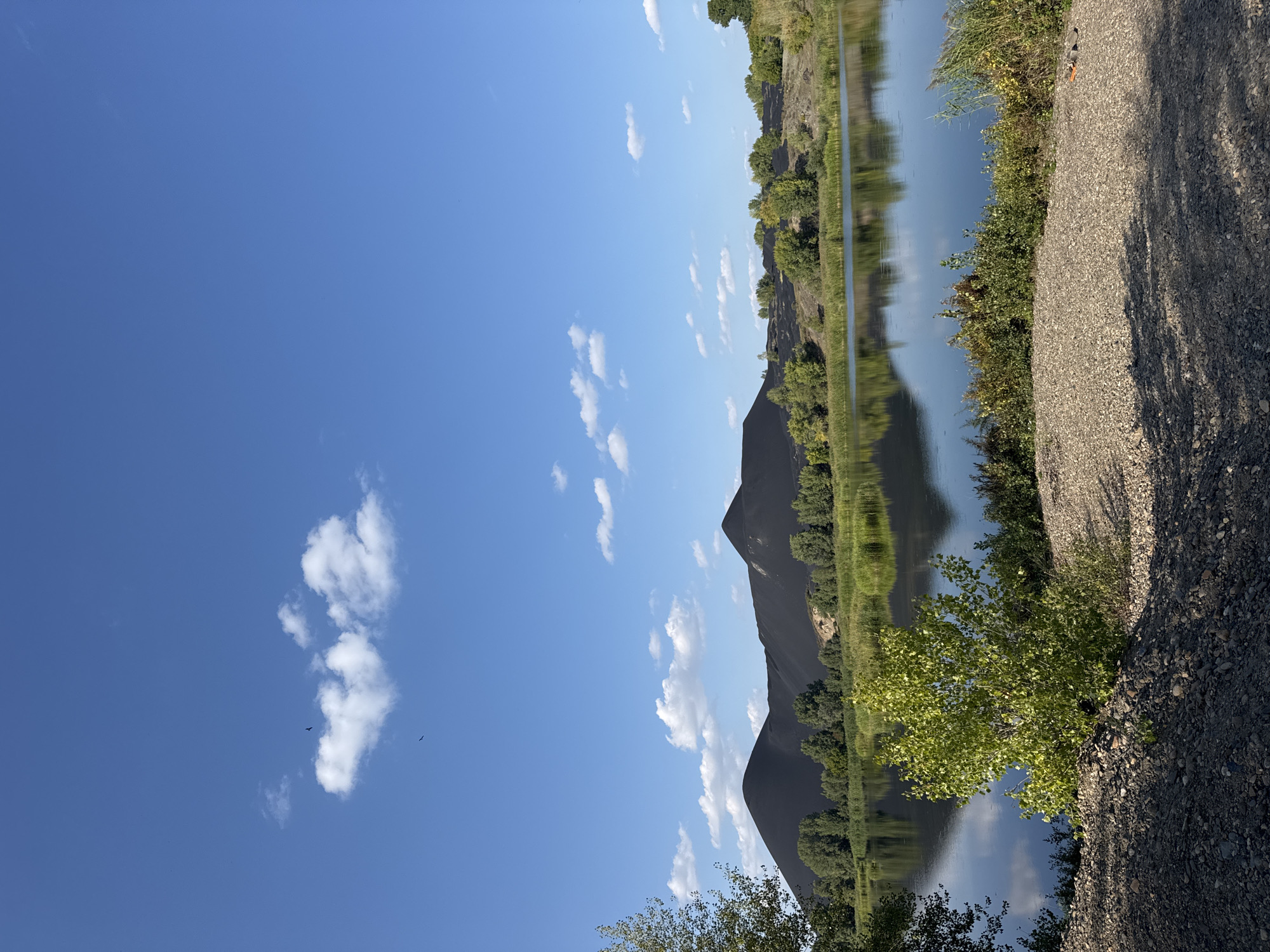 A wide shot captures a scenic view of a body of water with a clear blue sky above, dotted with a few wispy white clouds. In the distance, two large, dark, conical mounds dominate the horizon, likely slag heaps from industrial activity. Lush green trees and vegetation surround the water and partially obscure the base of the mounds. The water's surface is calm and reflects the sky and surrounding landscape, creating a mirror image. In the foreground, a rough, gravelly embankment leads down to the water's edge, with some green foliage growing from it. The overall impression is one of a natural landscape juxtaposed with signs of industry, likely located in the city of Glubokoe, Kazakhstan. There are no people or animals visible in the image.