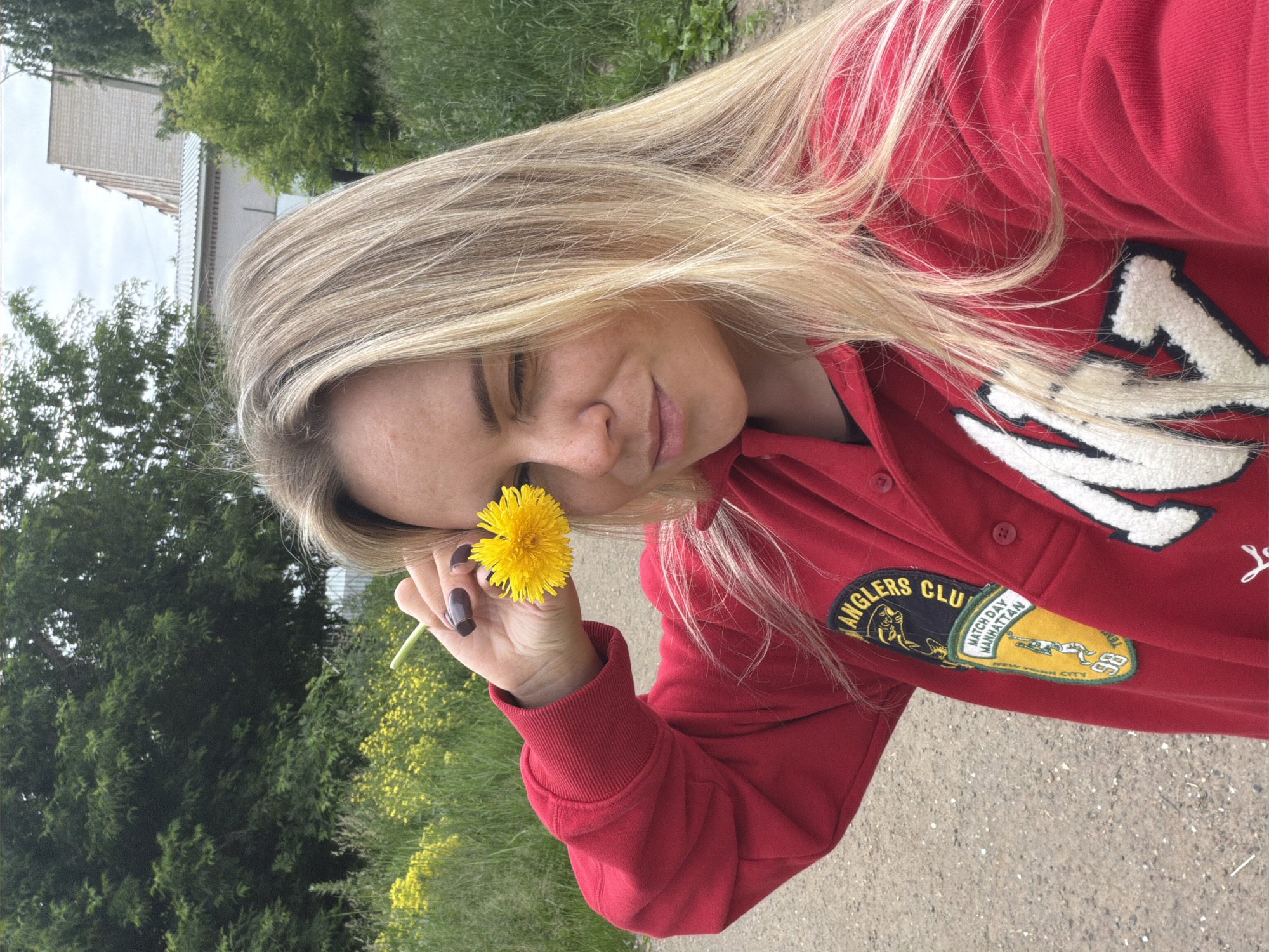 The image shows a young woman with blonde hair wearing a red sweatshirt. She is holding a yellow dandelion in front of her right eye, winking. The background consists of greenery, yellow flowers, a dirt path, and buildings. The scene is likely outdoors in a semi-rural or suburban area. Without additional information or context specific to Votkinsk, Russia, it is impossible to determine the exact location within the city.