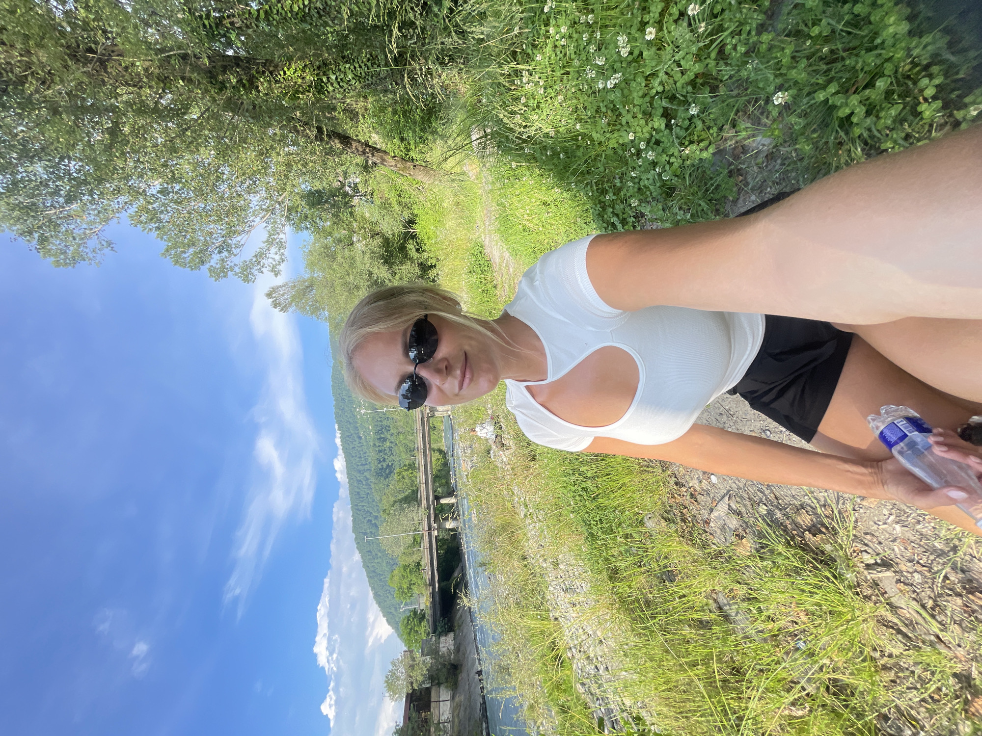 A woman with blonde hair, wearing sunglasses, a white cut-out top, and black shorts, is taking a selfie outdoors. She is holding a clear plastic bottle of water in her right hand. The woman is seated on a bank of rocks and grass, with a river flowing behind her. A bridge spans the river in the middle ground, and a lush, green mountainous landscape is visible in the background. The sky is blue with scattered white clouds. The location is the city of Sochi, Russia.