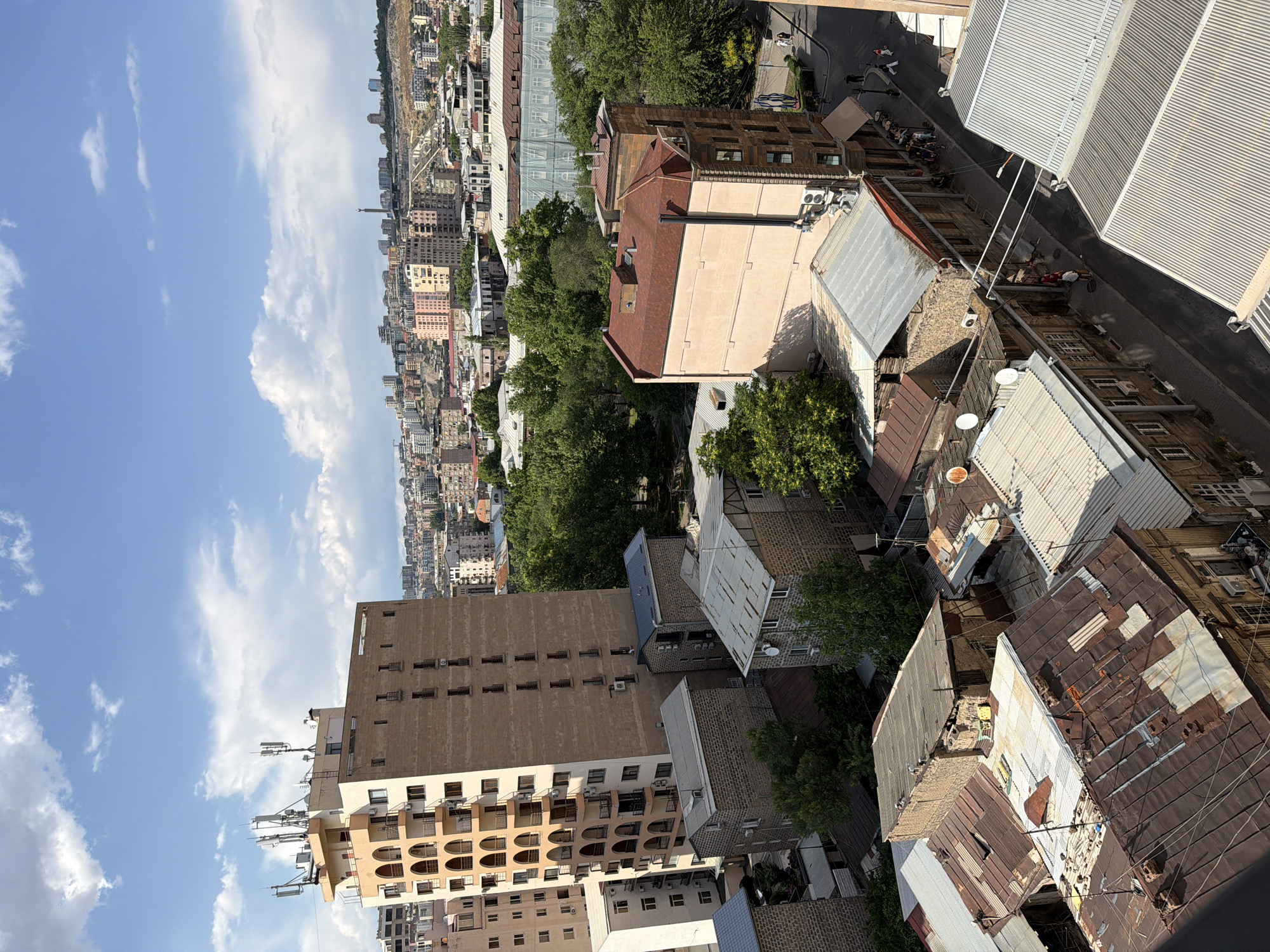 This is an aerial shot of a city street in Yerevan, Armenia, with a clear blue sky dotted with white clouds above. The foreground features a cluster of residential buildings, some with dilapidated, rusty corrugated metal roofs, suggesting an older or less maintained section of the city. In the mid-ground, a more modern, light-colored apartment building with arched balconies stands tall. Beyond this, a dense cityscape of multi-story buildings, some with red-tiled roofs, extends into the distance. Trees are interspersed throughout the urban landscape, particularly in the mid-ground, providing pockets of green. A street runs through the scene, with a few pedestrians visible, and a parked car on the right side. The overall impression is one of a densely populated urban environment with a mix of architectural styles and varying states of repair.