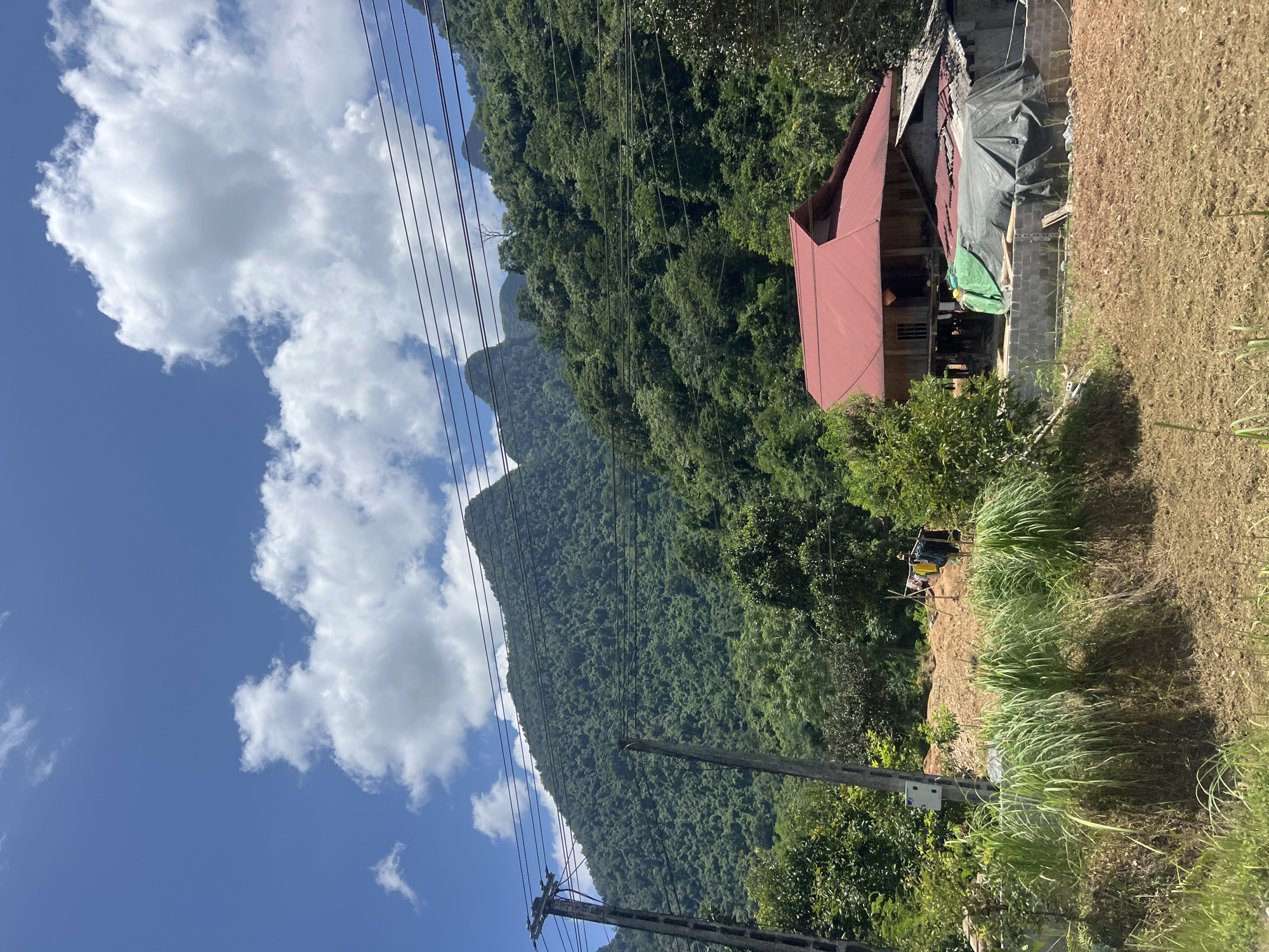 The image depicts a rural scene in Thuận Hòa Commune, Vietnam, characterized by lush green mountains under a blue sky with scattered white clouds. In the foreground, there is a cultivated field with brown soil and tall, green grass. A house with a red roof, built on stilts, is visible to the right, with another structure partially covered by a green tarp. Electrical poles and wires are present, traversing the landscape. The overall impression is one of a tranquil, mountainous countryside.