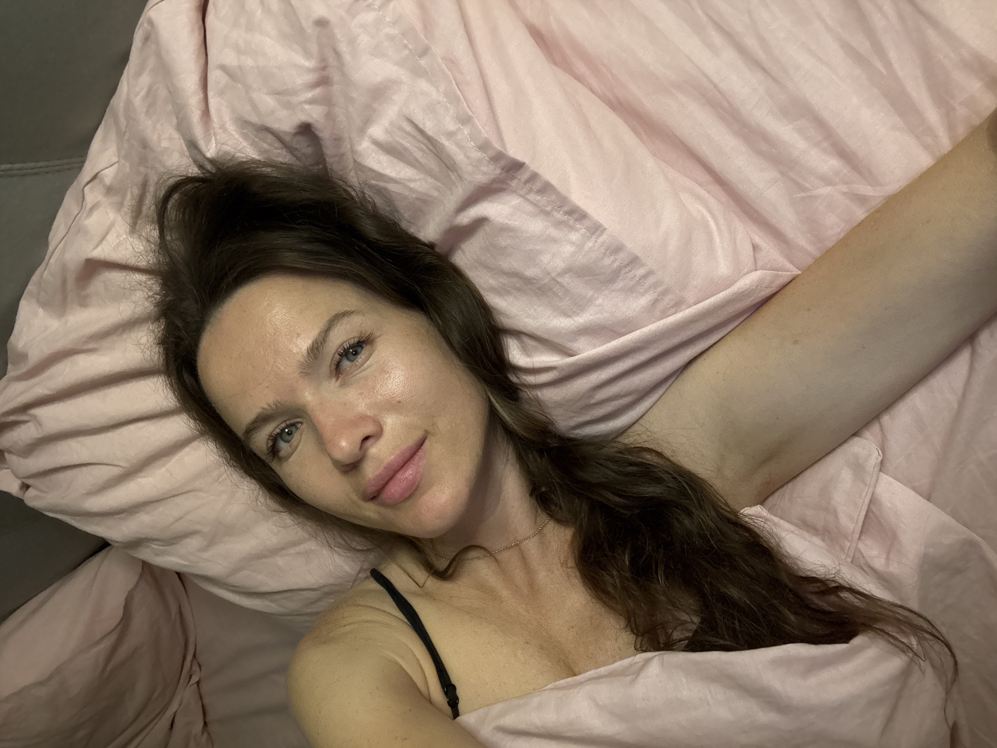 The image shows a woman lying in bed. She is in a supine position, with her head resting on a pillow. Her eyes are open, and she is looking at the camera. Her hair is dark brown and is spread across the pillow. She is wearing a black bra strap. The bed is covered with a pink duvet. The background appears to be a bedroom. There are no discernible landmarks or specific details that would indicate the location in Khimki, Russia.