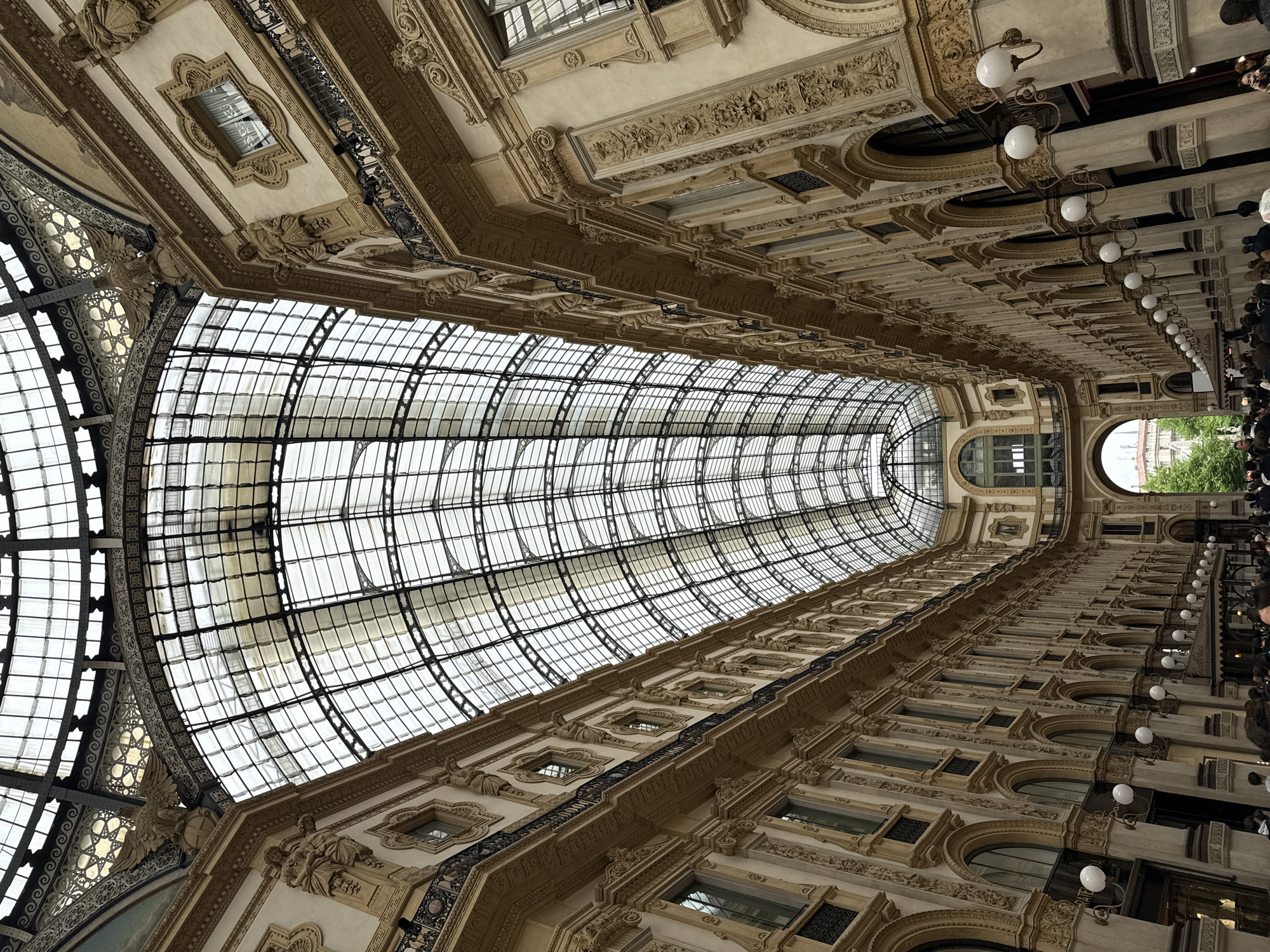 The image shows the interior of the Galleria Vittorio Emanuele II, a historic shopping gallery in Milan, Italy. The view is from below, looking up at the glass and iron roof structure and the ornate architectural details of the buildings that line the gallery.
The roof is a large arched structure made of glass panels held together by an iron framework. Light filters through the glass, illuminating the interior. The buildings on either side of the gallery are multi-storied with a classical design, featuring arches, columns, decorative moldings, and windows. There are also some people in the picture.