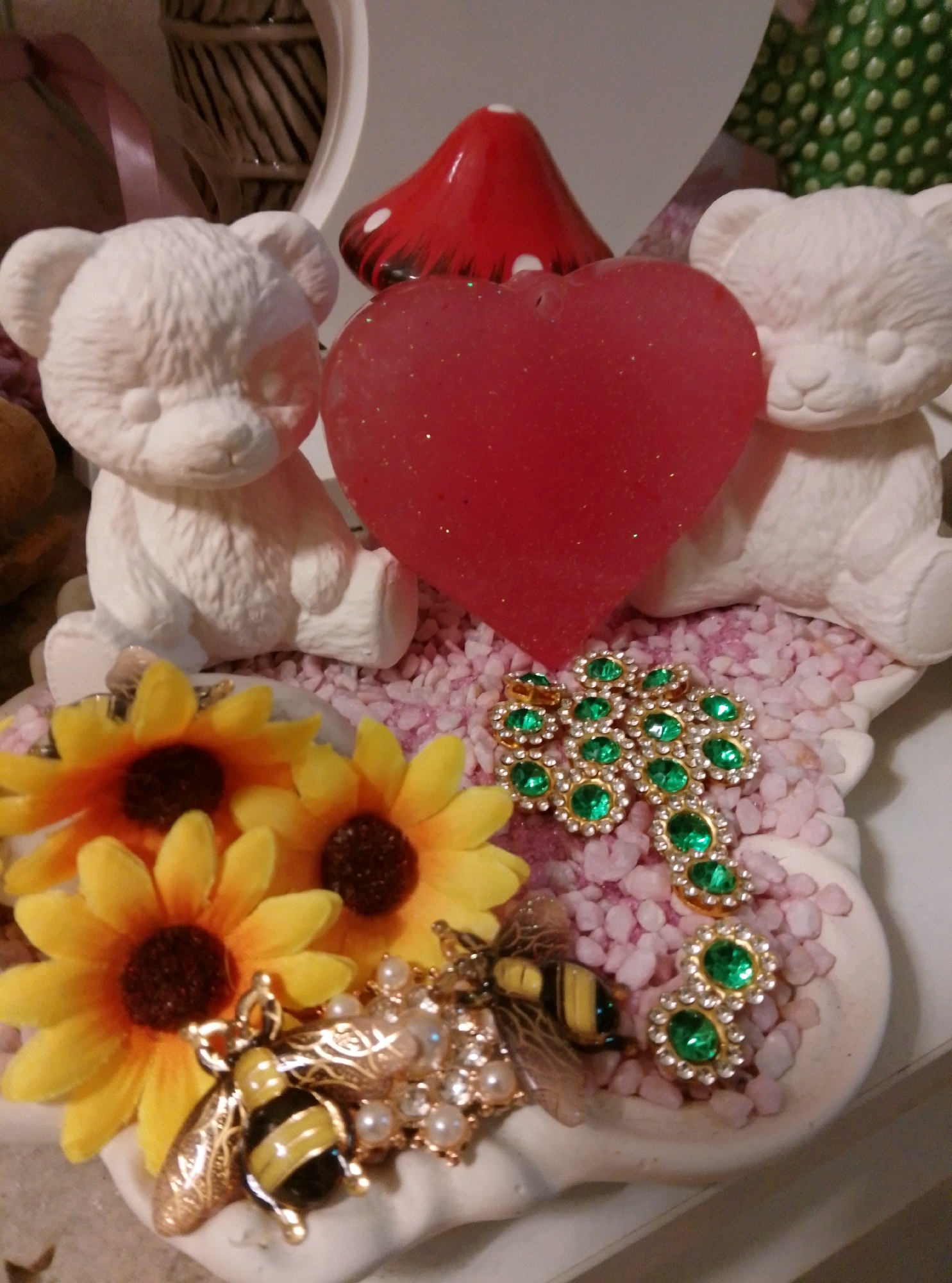 The image depicts a decorative display featuring two white ceramic teddy bears seated next to a large, translucent red heart. Behind them, a red mushroom figurine with white spots is visible. The display is set against a backdrop of woven material and a green fabric with a polka dot pattern. In the foreground, there are several yellow artificial sunflowers, a jeweled dragonfly-like embellishment with green stones and pearls, and a bee-shaped embellishment with black and yellow enamel and gold detailing. The entire arrangement rests on a surface covered with small pink pebbles. The context of Wedel, Germany is not discernible from the visual information provided in the image.