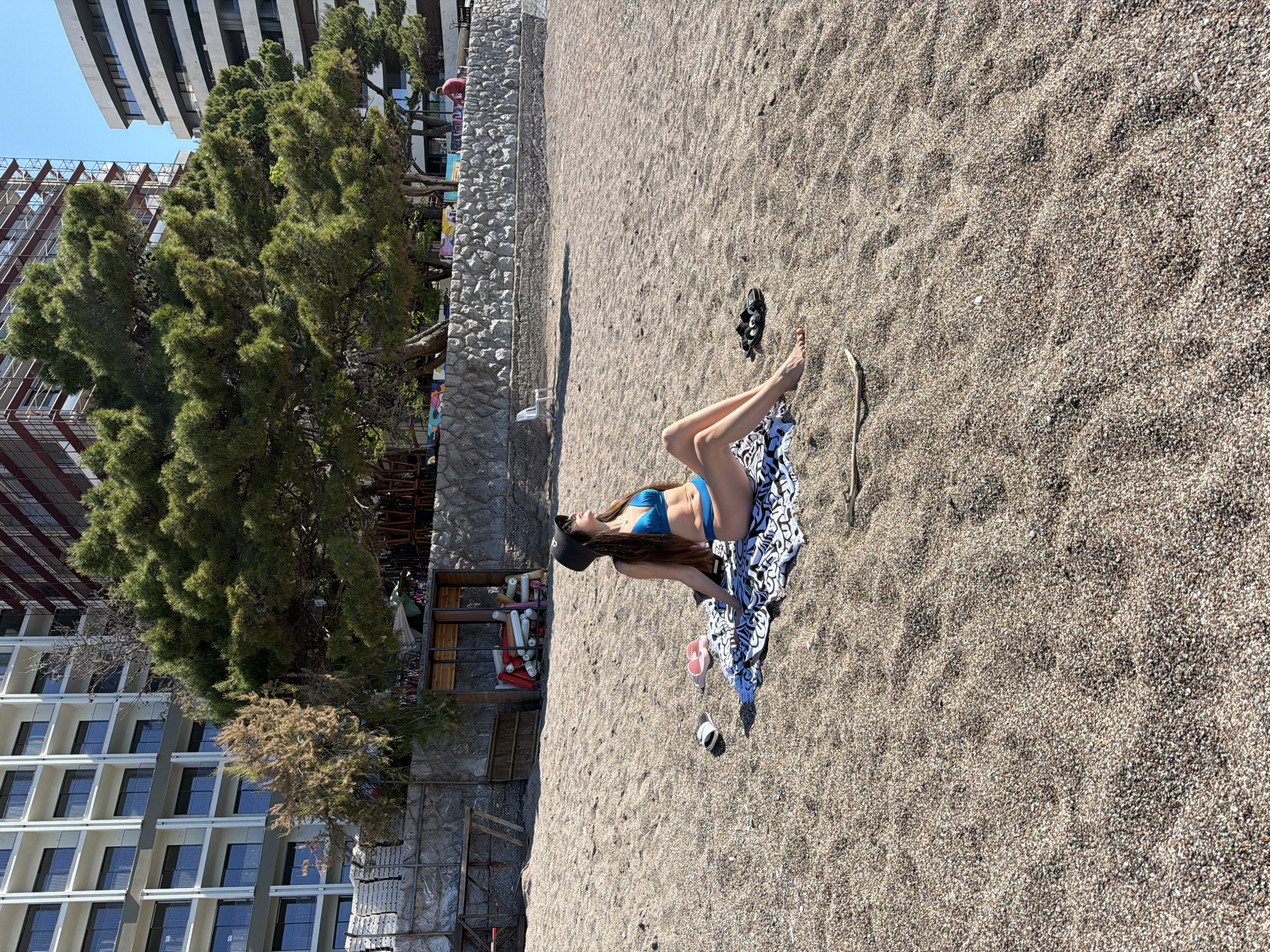 A high-angle, correctly oriented photograph captures a woman relaxing on a sun-drenched pebble beach in Rafailovići, Montenegro. The woman, with long brown hair, is lying on her side in a blue bikini top and a black baseball cap, facing towards the upper left of the frame. Her legs are partially covered by a black and white patterned beach towel. Nearby, a pair of black flip-flops and another pair of pink flip-flops are visible on the coarse, light-colored pebbles. A slender, light-colored stick or piece of driftwood lies on the sand near her feet.
The beach itself is composed entirely of small, smooth pebbles, extending from the foreground into the mid-ground. In the background, a low stone retaining wall runs horizontally, separating the beach from an area with lush green coniferous trees. Behind the trees, several tall, modern buildings are visible, suggesting a coastal resort or urban setting. One building on the left features a distinctive pattern of windows and balconies, while another appears to be under construction with exposed metal framing. The sky is clear and bright blue, indicating sunny weather. The strong sunlight casts distinct shadows, suggesting it is midday or early afternoon. The overall scene depicts a peaceful moment of relaxation by the sea.