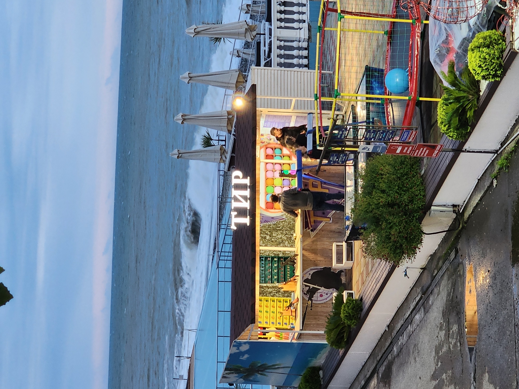 The image captures a seaside scene, likely in Sochi, Russia, evidenced by the Cyrillic text "ТИР" (shooting range) on a vertical sign. Turbulent waves crash on the shore, while a row of closed beach umbrellas suggests unfavorable weather. On a slightly elevated structure, what appears to be a shooting range or amusement stall is visible. Two people are present, one seated and another standing within the stall. The stall itself is brightly colored, decorated with various targets and designs. Adjacent to it, a colorful play structure with netting and a blue ball can be seen. Plants and greenery are strategically placed around the stall and play area, adding to the lively atmosphere. The area next to the structure is paved, and the image is taken from an elevated perspective, giving a comprehensive view of the seaside setting and the attractions present.