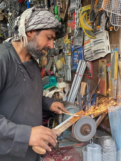 In Kandahar, Afghanistan, a man with a beard and a patterned head covering sharpens a machete. He is wearing a dark garment. The sharpening wheel spins rapidly, generating sparks. The scene is cluttered with tools and hardware, hanging on the wall. Chains, knives, and other metal implements are visible in the background.