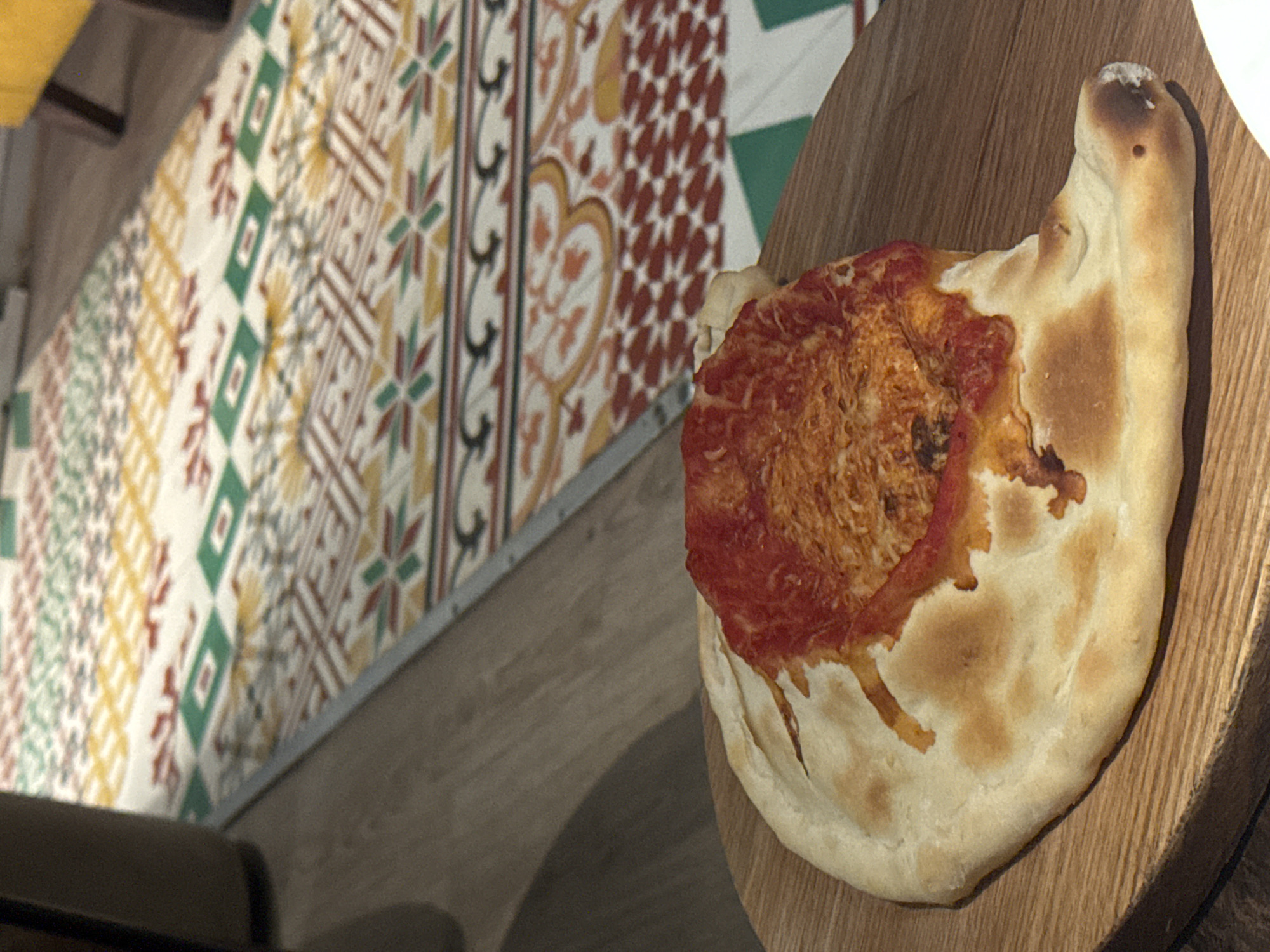 A calzone pizza rests on a wooden board. The pizza has a tomato sauce and cheese filling. In the background, a patterned carpet is visible. The scene is indoors.