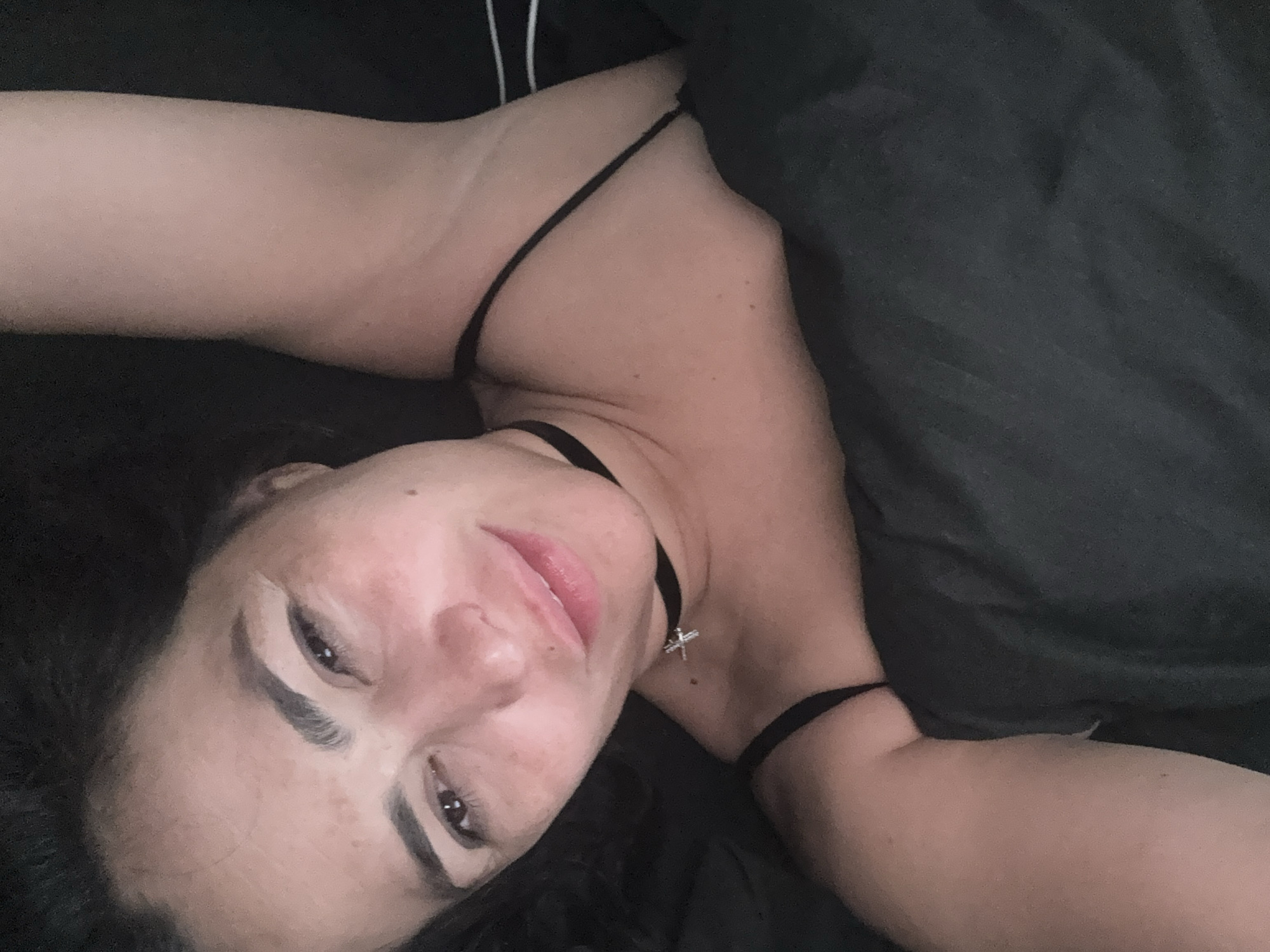A woman is lying in a bed. She is wearing a black spaghetti strap top and a black choker with a cross pendant. A dark gray blanket covers part of her body. The environment is dark. There is no information to confirm that this image was taken in the city of Chaikovsky, Russia.