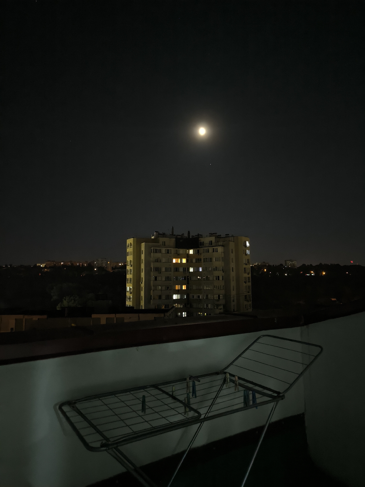 The image depicts a night scene in Chișinău, Moldova. In the foreground, a clothes drying rack is positioned on a balcony. Beyond the balcony, a multi-story apartment building is visible, with several windows illuminated by artificial light. The building appears to be residential. In the background, other buildings and the dark silhouettes of trees suggest a cityscape. The sky is dark, with the moon prominently visible, casting a soft glow. A few faint stars are also discernible. The overall impression is one of a quiet urban night.