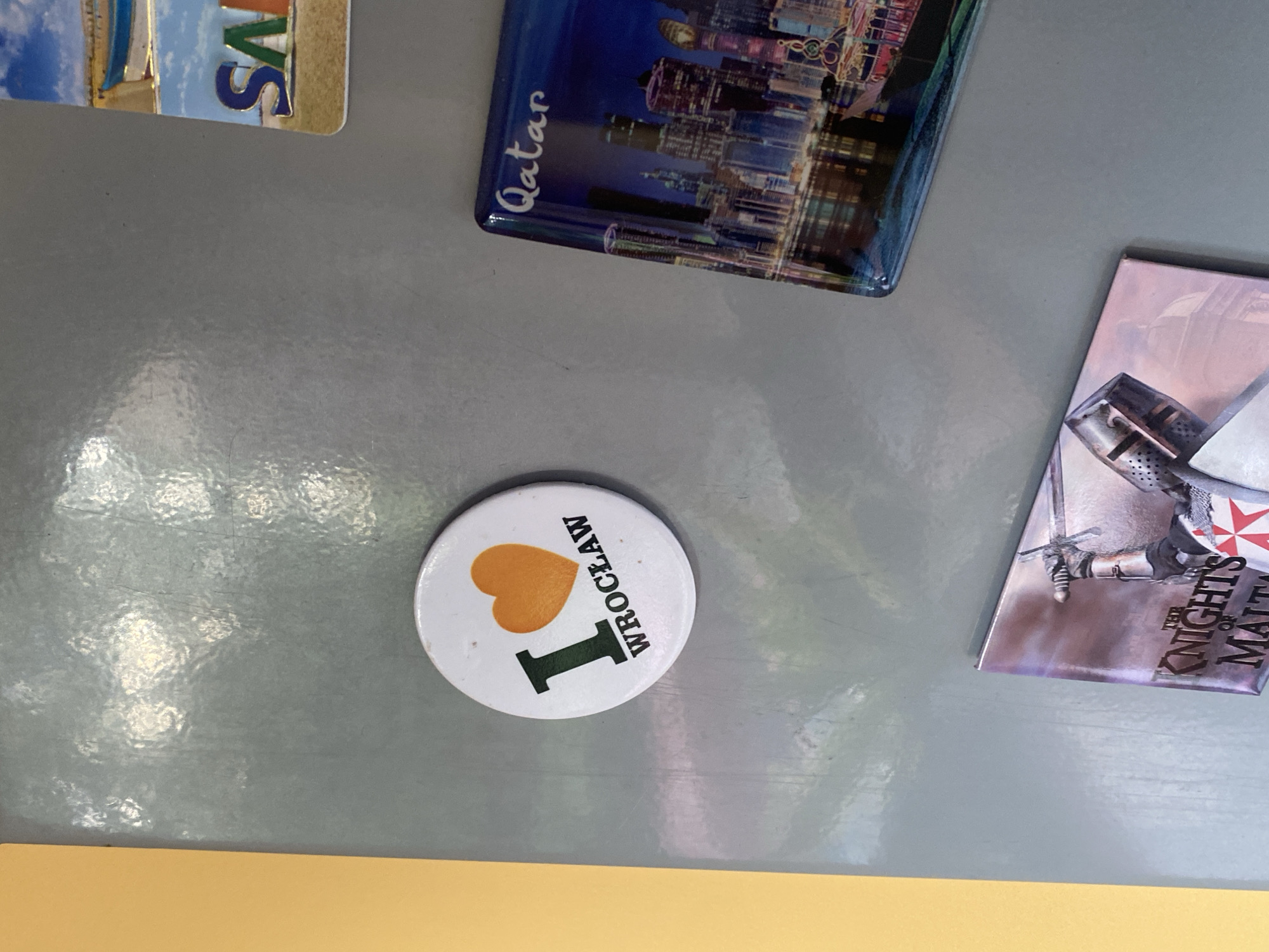 A close-up, eye-level shot shows three magnets on a gray, textured refrigerator door, to the left of a bright yellow vertical surface. The topmost magnet features a landscape scene with a blue boat on a sandy beach under a bright sky. The middle magnet is a round, white badge with black text that reads "I ❤️ WROCLAW". Below that, a magnet depicts a knight in armor with a sword, part of a "THE KNIGHTS OF MALTA" design. To the right of the badge magnet, a rectangular magnet displays a nighttime cityscape with illuminated skyscrapers and the word "Qatar" in white script. The lighting is bright, casting reflections on the refrigerator surface.