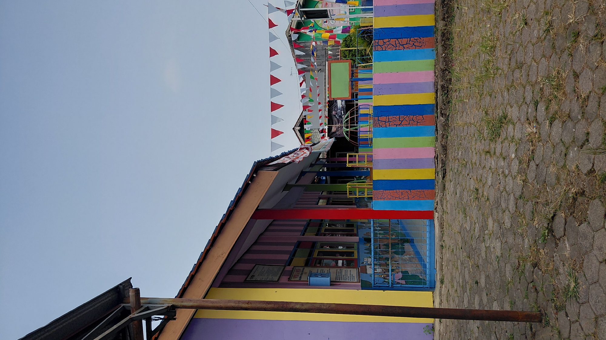 An outdoor view of a colorful, low-rise building with a covered walkway, likely a school or community center, is shown under a clear sky. The building features brightly painted walls in shades of purple, yellow, red, green, and orange. A blue metal gate is visible in front of the building. A colorful fence, decorated with vertical stripes of various colors, runs along the front of the property. Flags are strung across the top of the fence. The ground in front of the building is paved with hexagonal concrete blocks. The scene appears to be in a semi-urban or rural setting, possibly in the city of Rejasari, Indonesia, given the context. No people are present in the image.