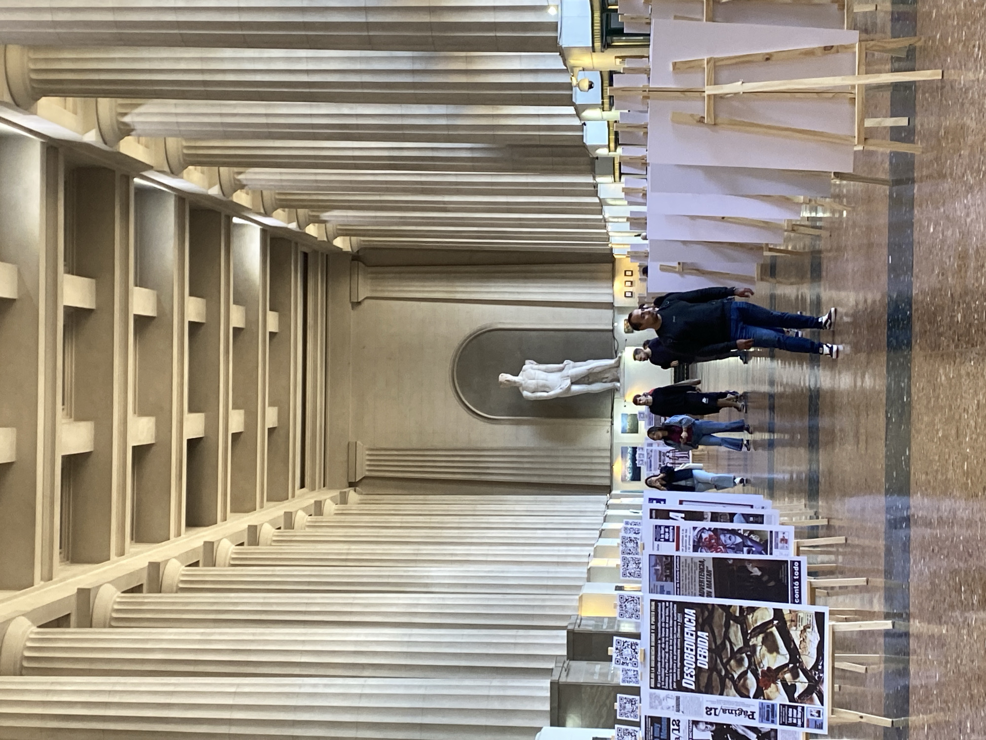 The image captures a long corridor within a building, likely a museum or cultural center, in Buenos Aires, Argentina. The architectural style is characterized by numerous tall, neoclassical columns lining the left side, creating a colonnade effect. Near the end of the corridor, a white statue of a nude male figure stands in a recessed alcove.
On the right side of the corridor, several wooden easels are arranged, displaying various posters and sheets. Some of these posters have visible text including "DESOBEDIENCIA DEBIDA" and "Página/12," along with QR codes. Further down the corridor, more easels hold blank white boards.
A group of people is present, walking along the corridor. They appear to be viewing the posters. The floor is a patterned stone or tile. Overhead lighting is visible towards the end of the corridor. The scene suggests an exhibition or display within a historical building in Buenos Aires.