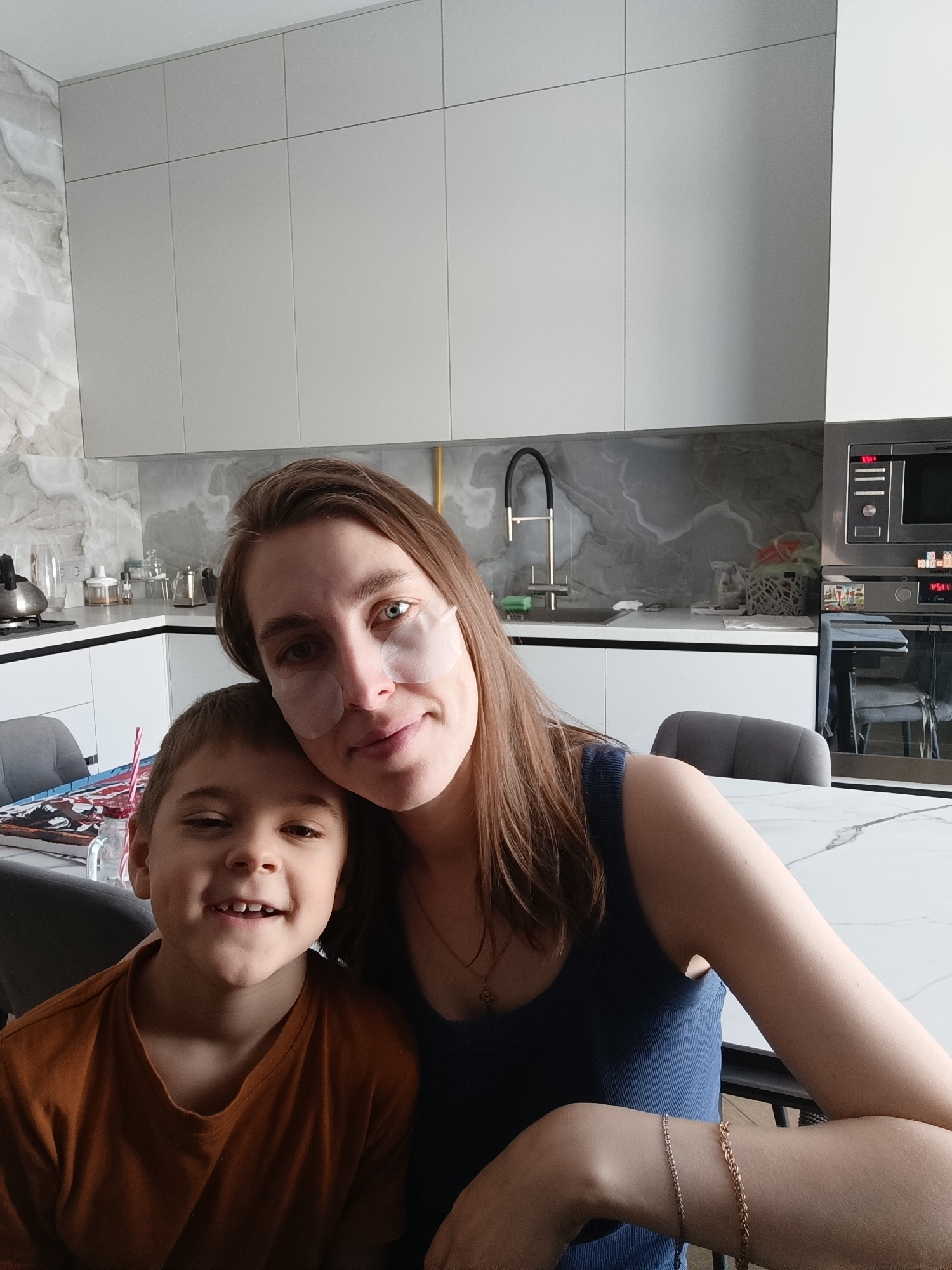 A woman and a boy pose closely together in what appears to be a kitchen setting. The woman, fair-skinned with light brown hair, is wearing eye patches on her face and a blue top. She has a bracelet on her right wrist. The boy, who is wearing a brown shirt, is smiling.
Behind them, the kitchen features white cabinets, a marble-patterned countertop, a faucet, a built-in microwave/oven unit, and dining chairs.
While the precise location in Chaikovsky, Russia, cannot be confirmed from the image alone, the photograph was geolocated there.