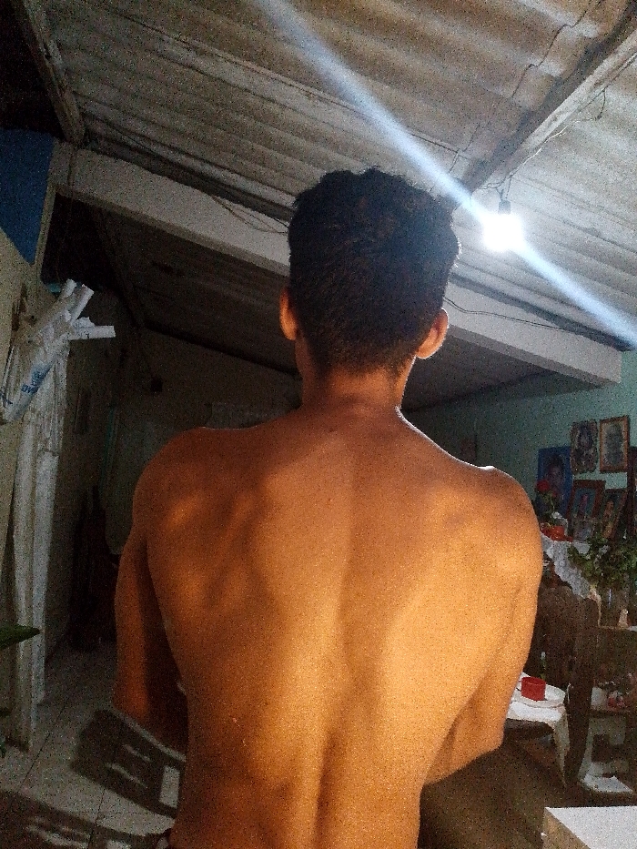 The image depicts a male individual, observed from behind, standing within an interior space in Juan Griego, Venezuela. The subject has short, dark, curly hair and is shirtless, revealing his muscular upper back and shoulders, illuminated by a direct light source. The overhead ceiling is corrugated, appearing to be unfinished or utilitarian, with exposed electrical wiring and a single bare light bulb hanging centrally, emitting a strong white light that creates prominent lens flares. To the subject's left, a light-colored wall features stacked or hanging white material and a darker recessed opening. To the right, a greenish-blue wall displays multiple framed pictures or religious artwork above a white-draped surface, possibly a small table or altar, on which a red object is visible. The floor in the lower left foreground appears to be tiled or concrete. The scene is characterized by artificial lighting.