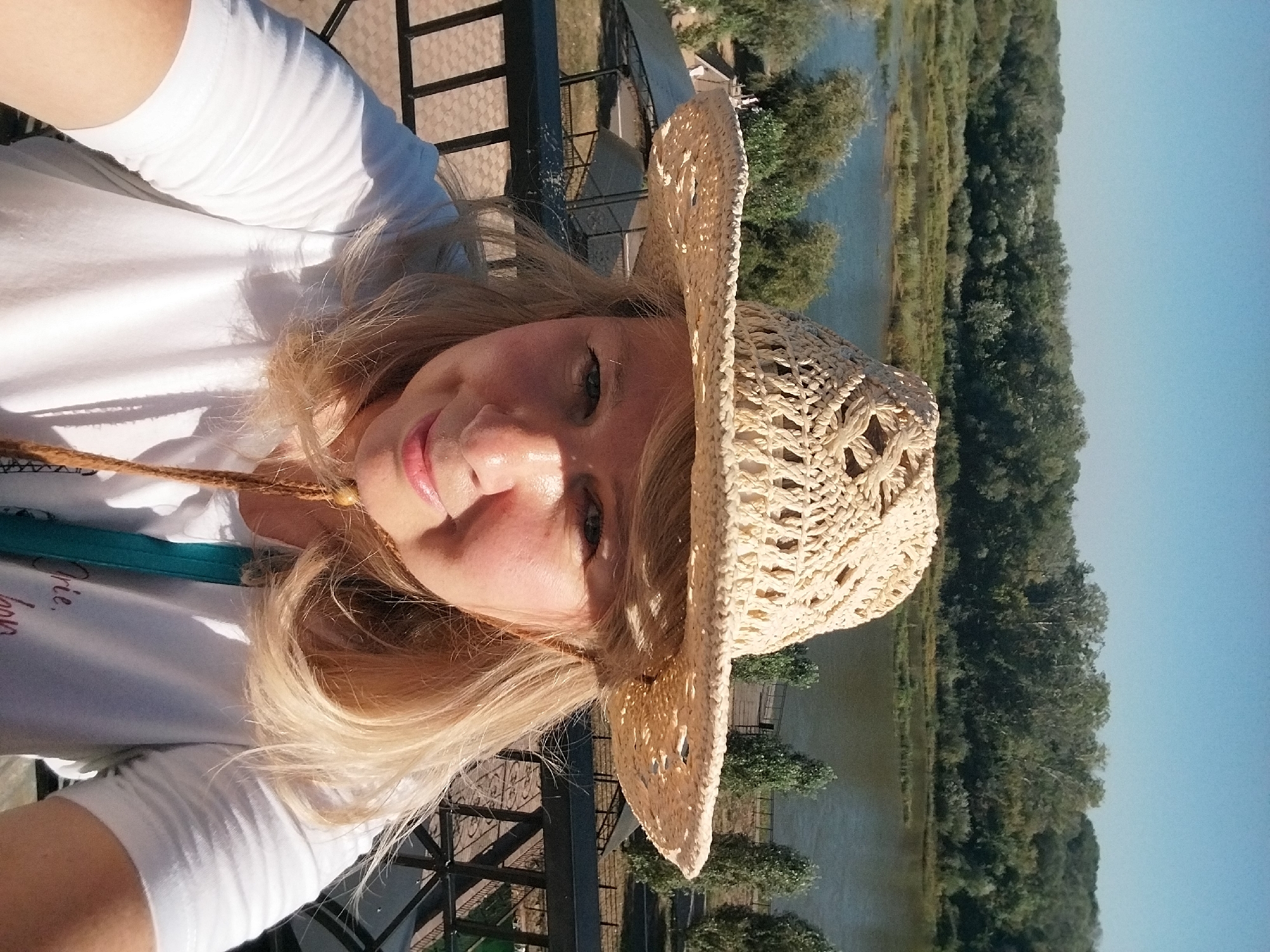 A woman wearing a straw hat and a white t-shirt is taking a selfie outdoors. She has blonde hair and is smiling. In the background, there is a body of water, trees, and a clear blue sky. The location is not specified as Orenburg, Russia.