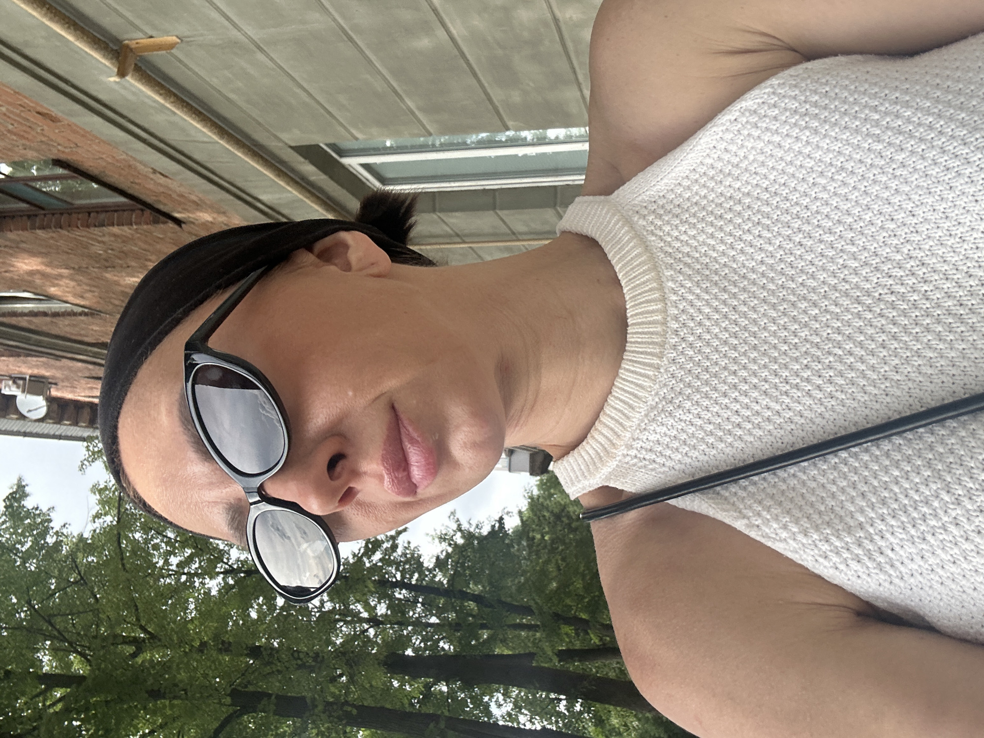 A close-up, waist-up, selfie-style shot shows a woman wearing a white, knitted halter top and black sunglasses. She has dark hair pulled back and is outdoors with trees and a brick building in the background. The building appears to be in an urban environment. The lighting suggests it is daytime. The woman's face is turned to her left, with her mouth slightly parted. The overall impression is a casual, candid moment captured in an outdoor setting.