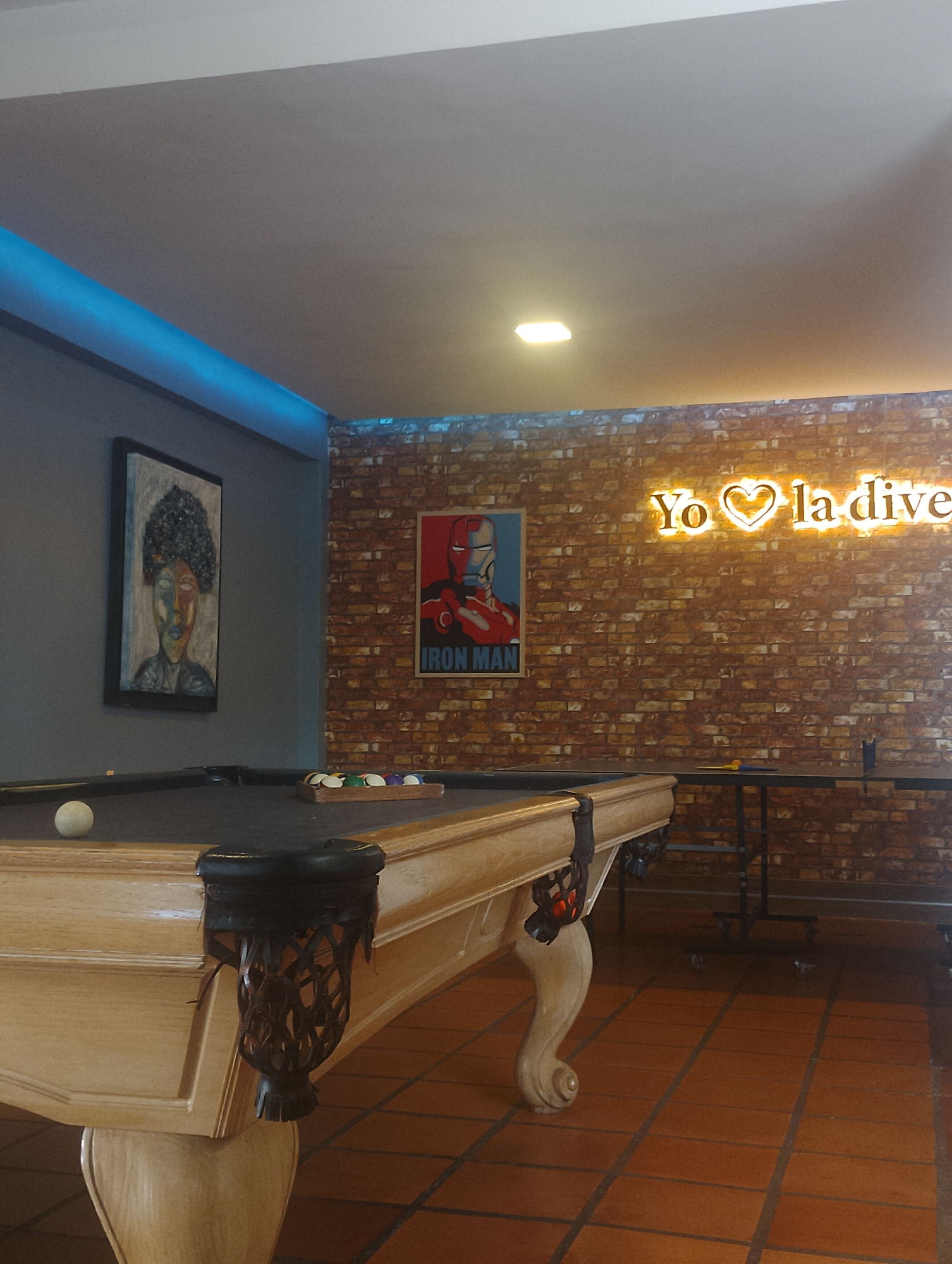 The image depicts an indoor recreational space. A pool table dominates the foreground, positioned on a red tile floor. The table is light-colored with black pockets and a set of billiard balls arranged in a wooden rack. On the left wall hangs a framed portrait of a person with dark hair and colorful face.
The back wall, constructed of red brick, features two decorations. On the left is an "Iron Man" poster, and on the right is a neon sign reading "Yo❤️ la dive." To the right of the pool table is a black table. The overhead lighting and a blue light strip around the room's perimeter contribute to the overall illumination. The room is likely a game room or entertainment area. The location is Porlamar, Venezuela. There are no people visible in the frame.