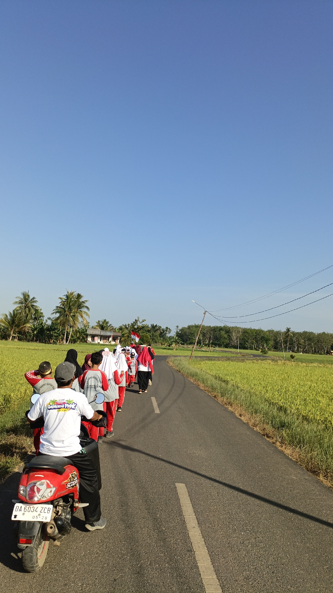 In Batarang, Indonesia, a group of people, likely students and adults, walk along a paved road lined with lush green rice fields. The sky is a clear, vibrant blue. A man in a white shirt and black pants rides a red scooter in the foreground, facing away from the camera. He is following the group. Some of the walkers are wearing red and white attire, and several of the women are wearing hijabs. One person in the group is carrying a flag. In the distance, there are palm trees and a small building. Overhead, power lines stretch across the scene. The overall impression is of a peaceful, outdoor activity taking place in a rural setting.