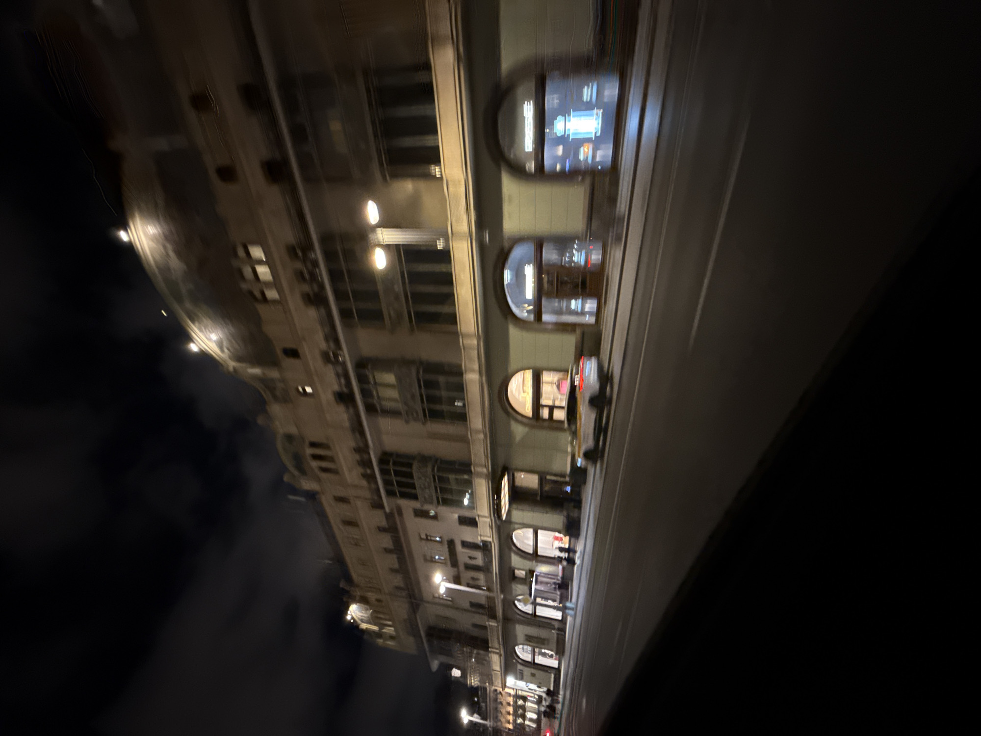 This is a photograph taken at night in Moscow, Russia, featuring the iconic GUM department store on Red Square. The image is slightly blurry due to motion, likely from a moving vehicle.
The main subject is the grand facade of GUM, a historic building with a distinctive glass roof (partially visible) and multiple stories. The architecture is detailed, with arched windows and ornate balconies. Lights illuminate the building's exterior, highlighting its classical design.
In front of the building, a street with white lane markings is visible, suggesting it is a public road. Several cars are present: a light-colored car, possibly a sedan, is parked or moving on the right side of the road, and a yellow and white taxi is visible further down the street. A few figures, appearing to be pedestrians, are seen walking on the sidewalk in front of the shops within GUM.
The sky above is dark with some cloudy formations, indicating nighttime. The overall atmosphere is urban and festive, with the illuminated architecture creating a sense of grandeur. The image captures a moment of city life at night, with the blend of historical architecture and modern activity.