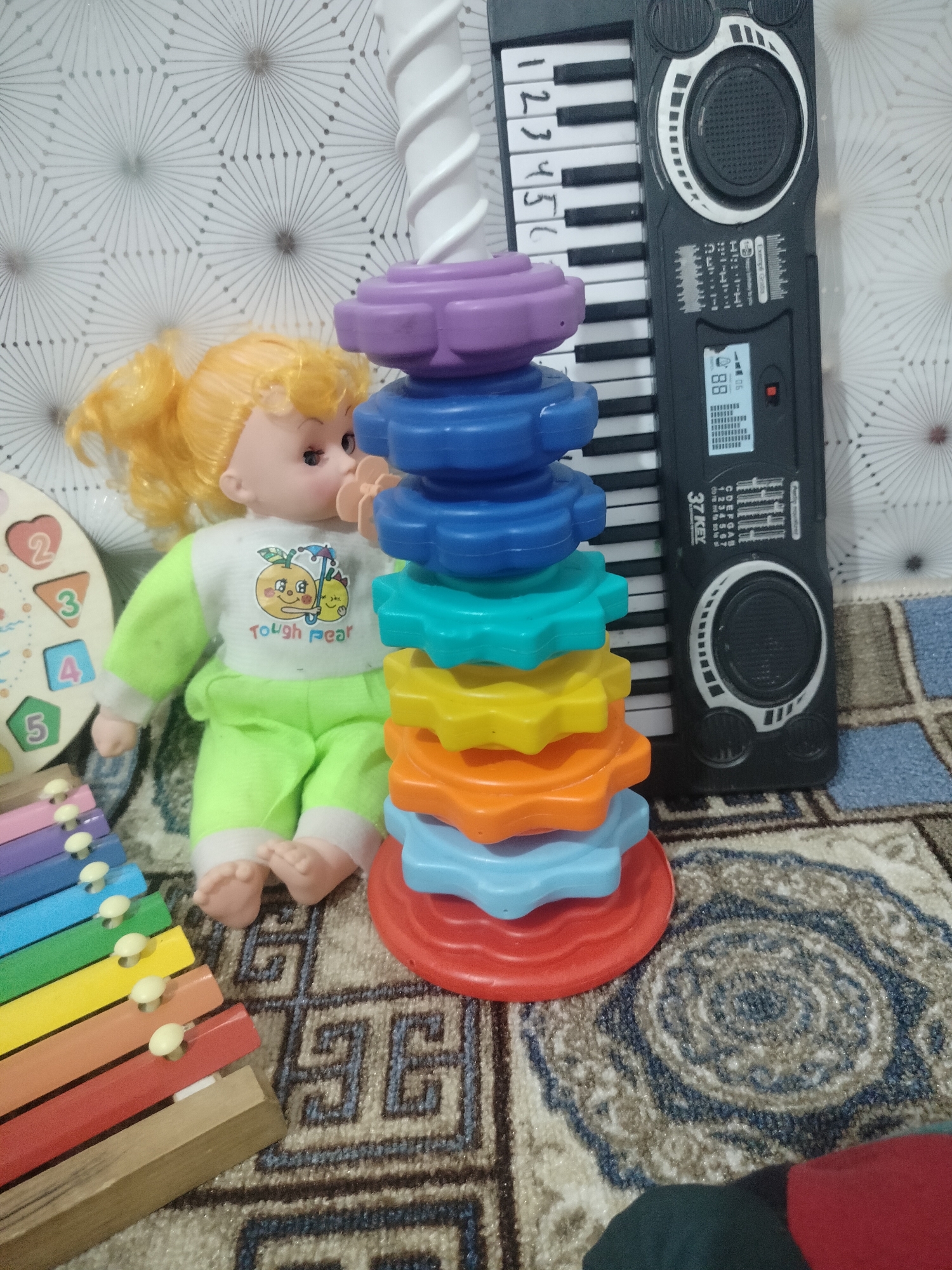The image shows a collection of children's toys on a patterned rug. In the foreground, a colorful xylophone is visible. To the right of the xylophone, a doll with blonde hair is seated, wearing a light green outfit. A stack of brightly colored stacking rings is placed next to the doll. Behind the stacking rings, a toy keyboard with black and white keys is positioned. The background features a textured, possibly wallpapered, surface with a geometric starburst pattern. The overall setting appears to be indoors, likely a child's play area. The location in the city of Cholpon, Kyrgyzstan, is not determinable from the visual information presented.