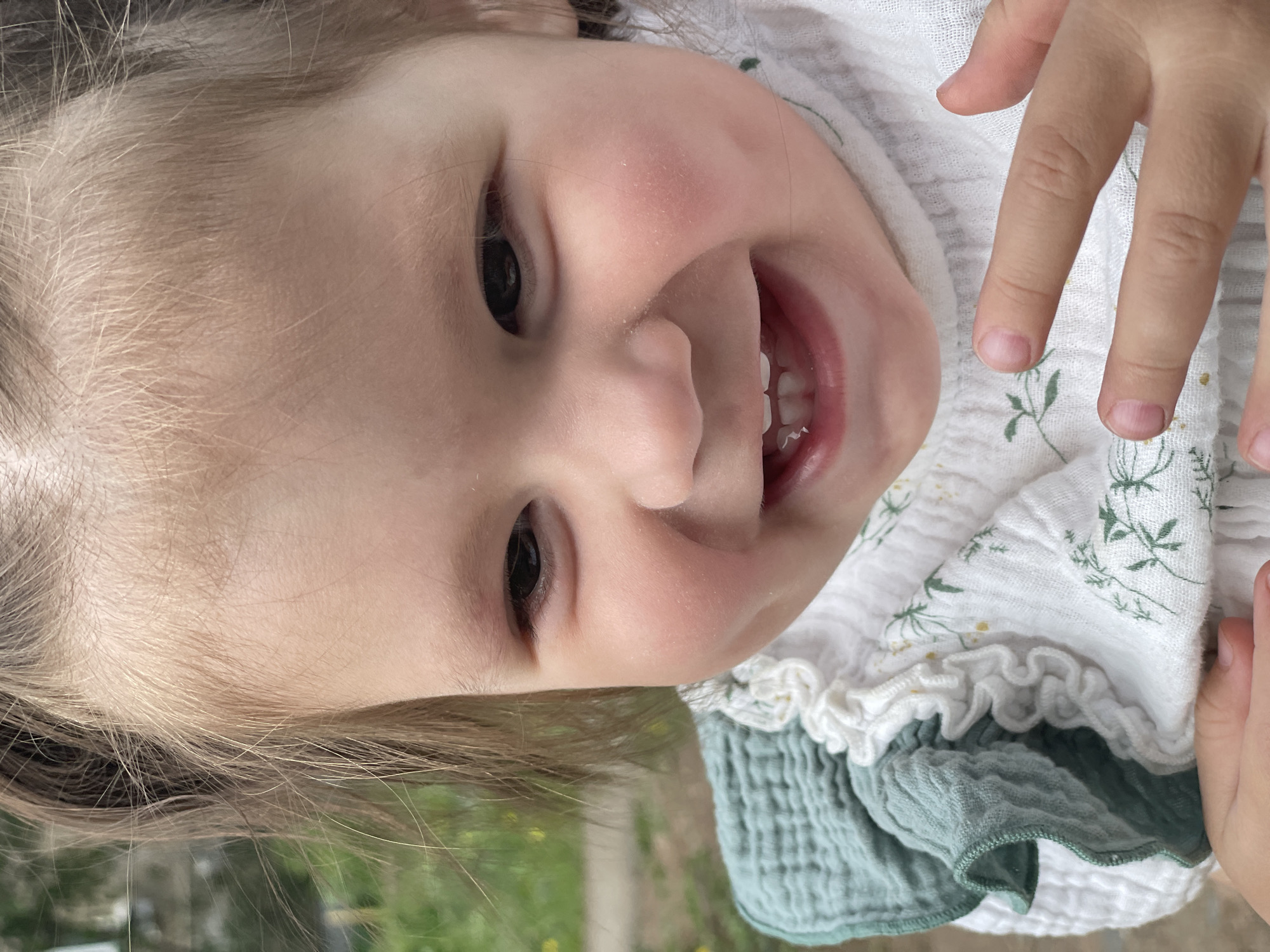 A close-up portrait of a young child with light skin and light blonde hair, smiling and showing their teeth. The child is wearing a white top with green floral embroidery and a ruffled collar. A green garment is visible beneath the white top. Part of the child's hand is visible at the upper right. The location is not identifiable.