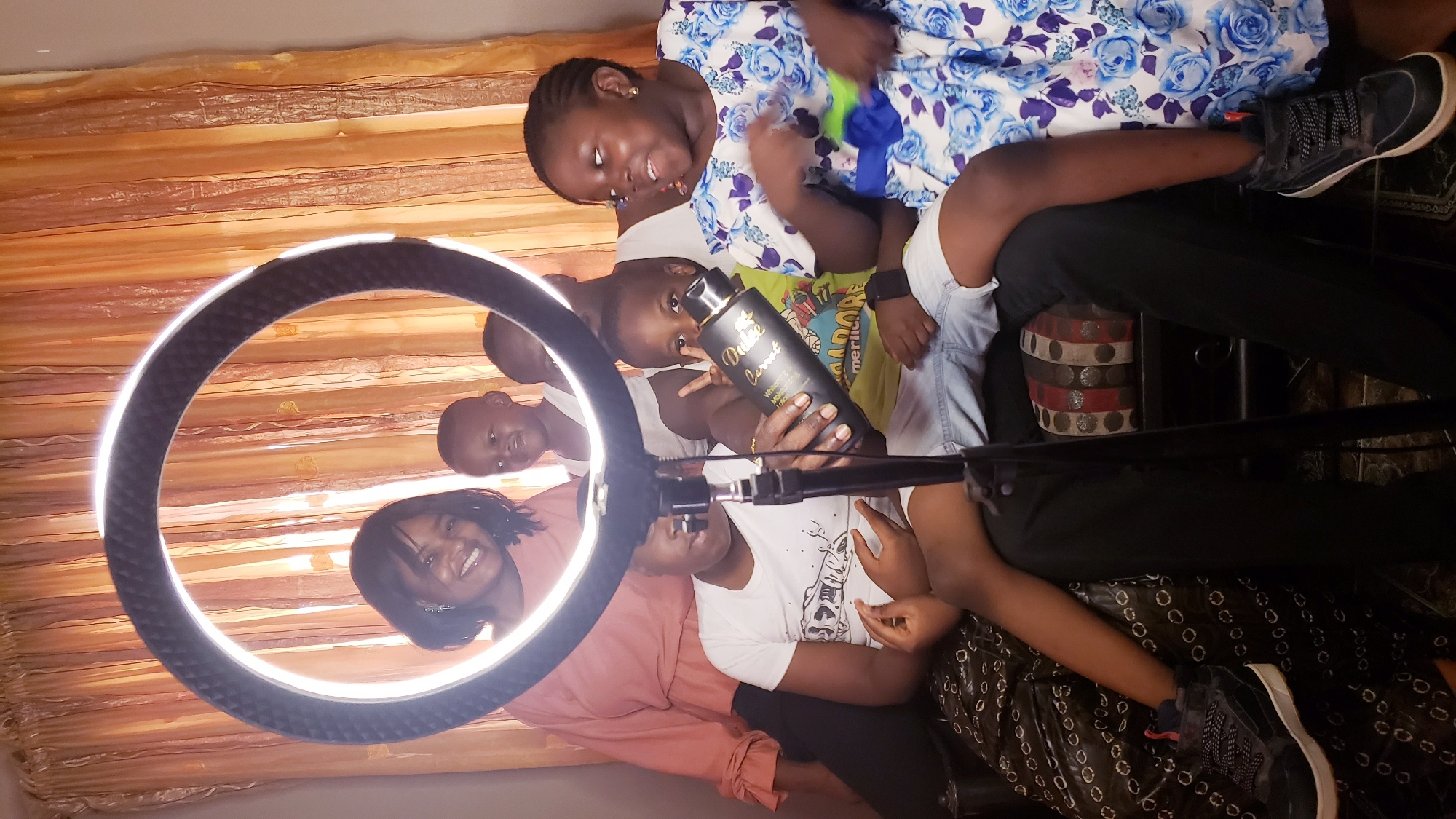 A ring light stands in front of a group of seven people in what appears to be an indoor setting in Sabon Tasha, Nigeria. An adult woman, positioned on the left of the ring light, smiles at the camera, dressed in a light pink top. To the right of her, two younger children look toward the viewer; one child is holding a black bottle labeled "Dubai Sweet." At the top of the frame, an older girl wears a floral patterned top and plays with a blue and green object. Beneath her, another young child wears a yellow t-shirt with cartoon imagery. The people are partially seated or leaning on a piece of dark furniture with striped decorative elements. The background features a peach-colored, semi-transparent curtain. A pair of black sneakers are seen at the edges of the frame, worn by someone seated.