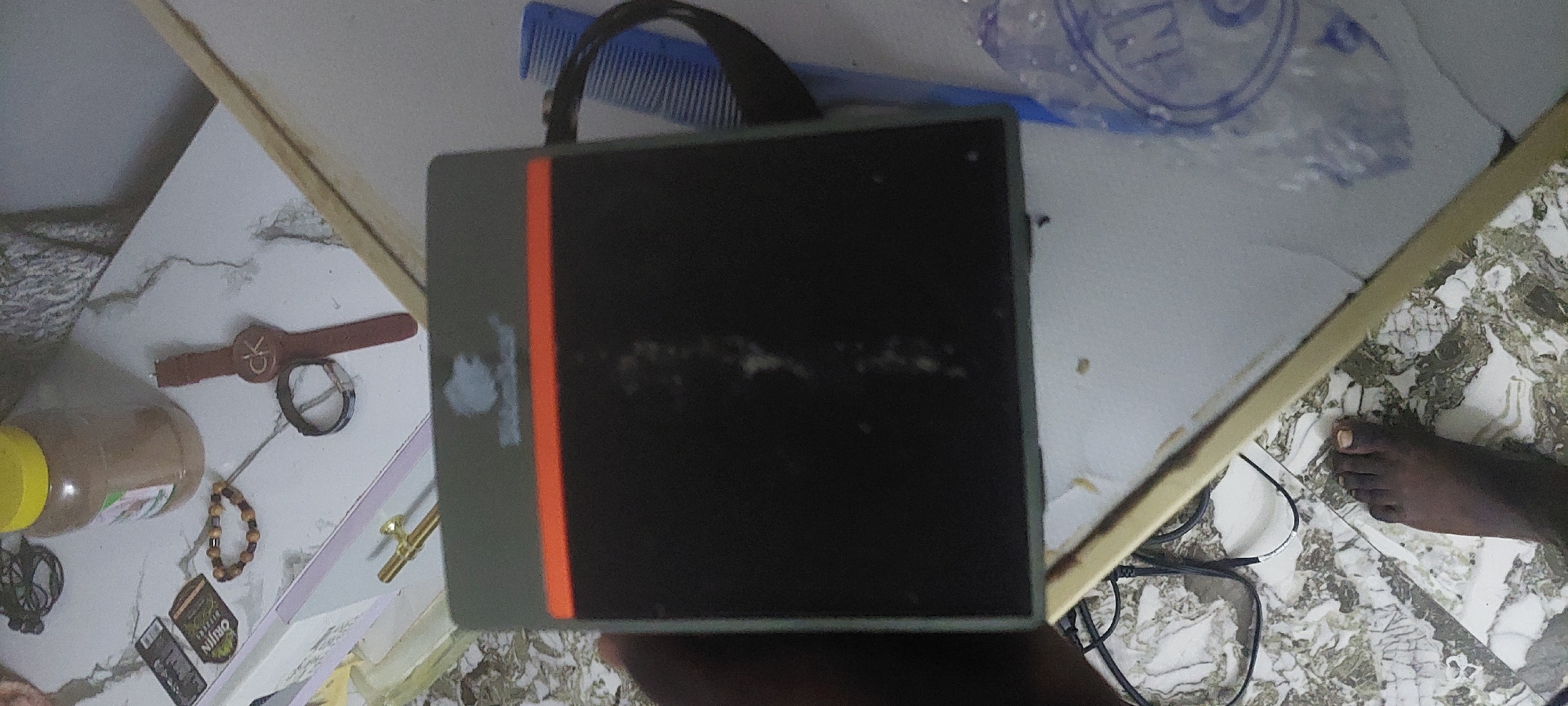 The image depicts a still life on a marble-patterned surface, likely a table or shelf, in what appears to be an indoor setting. In the foreground, a dark-skinned foot is visible, suggesting a person is present but mostly out of frame. To the right of the foot, a blue comb is partially visible, attached to a dark, box-like object with an orange stripe and a light-colored logo. Above and to the left of this object, a brown jar with a yellow lid is prominent, alongside a beaded bracelet and a watch with a "CK" logo. Various small items, including what look like cards or labels, are scattered on the surface. The background shows a textured wall and hints of other objects, possibly furniture. The overall impression is of a disorganized personal space. Based on the provided context, this scene is located in Jalingo, Nigeria.