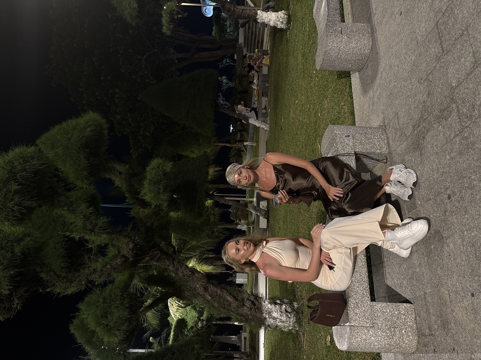 Two women pose for a photo at night. The first woman wears a dark brown satin dress and a headscarf. The second woman wears a light tan halter dress and pants. Both women wear white sneakers. They sit on a grey stone structure in front of a lawn. A dark brown purse sits on the stone next to the second woman. In the background, people sit at a table in a park. The photo was taken in Nha Trang Ward, Vietnam.