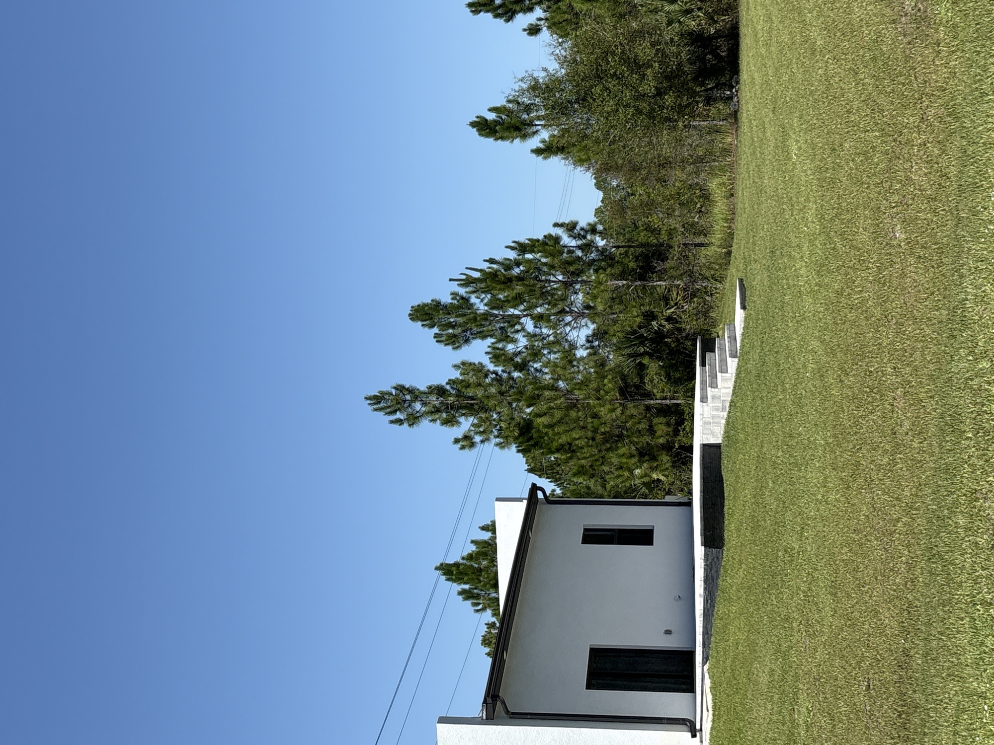 A modern white house with black trim and dark doors sits on a lush green lawn under a clear blue sky. The house has a simple, rectangular design with a flat roof and a few windows. A set of concrete steps leads up to a raised patio area in front of the house. Behind the house, a dense line of tall pine trees and other greenery forms a natural backdrop. Power lines stretch across the sky above the trees. The scene appears to be in a semi-rural or suburban area of Lehigh Acres, Florida, United States of America, characterized by open space, well-maintained landscaping, and a clear, sunny climate. There are no people or animals visible in the image.