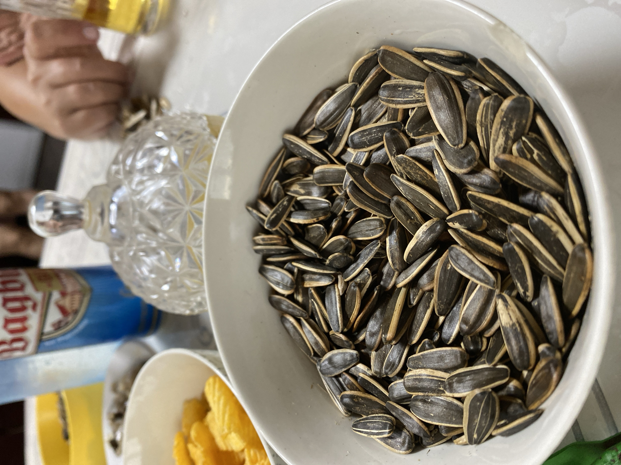 A close-up shot focuses on a white bowl filled with black and white striped sunflower seeds. The seeds are piled high in the bowl, with some spilling over the edge. In the background, out of focus, are other items on a white table. These include a blue can with red lettering that appears to say "Bagby", a decorative glass jar, and a person's hand reaching towards a small pile of sunflower seeds. A glass of golden liquid, possibly beer, is also visible to the right. The scene suggests a casual gathering or snack time. The image is taken from a slightly elevated angle. The location is not identifiable from the image.