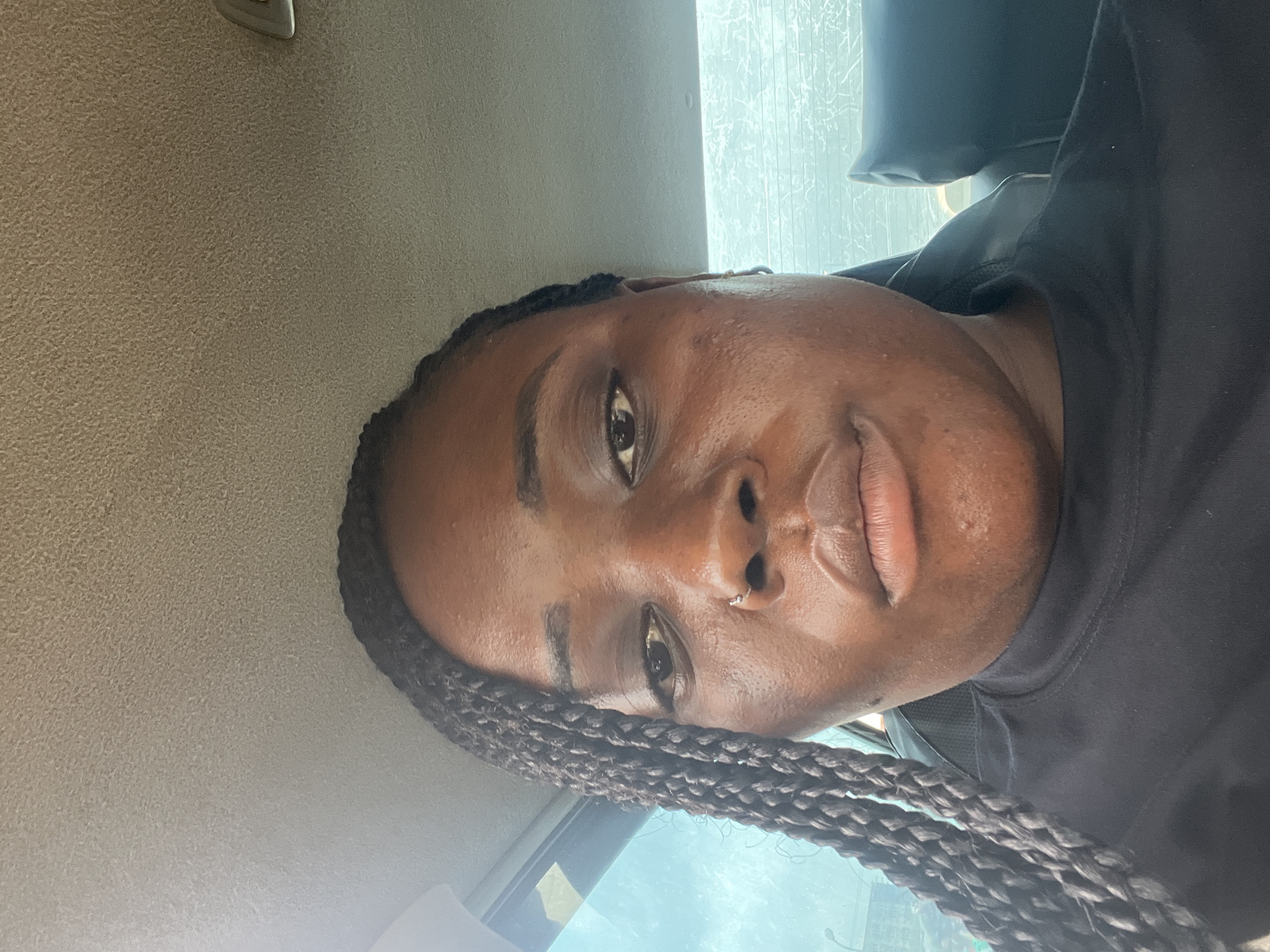 A close-up, tilted portrait shows a dark-skinned person with braided hair. The individual has a nose ring and is wearing a black shirt. The background suggests the interior of a vehicle. There is no information to indicate the location is Abuja, Nigeria.