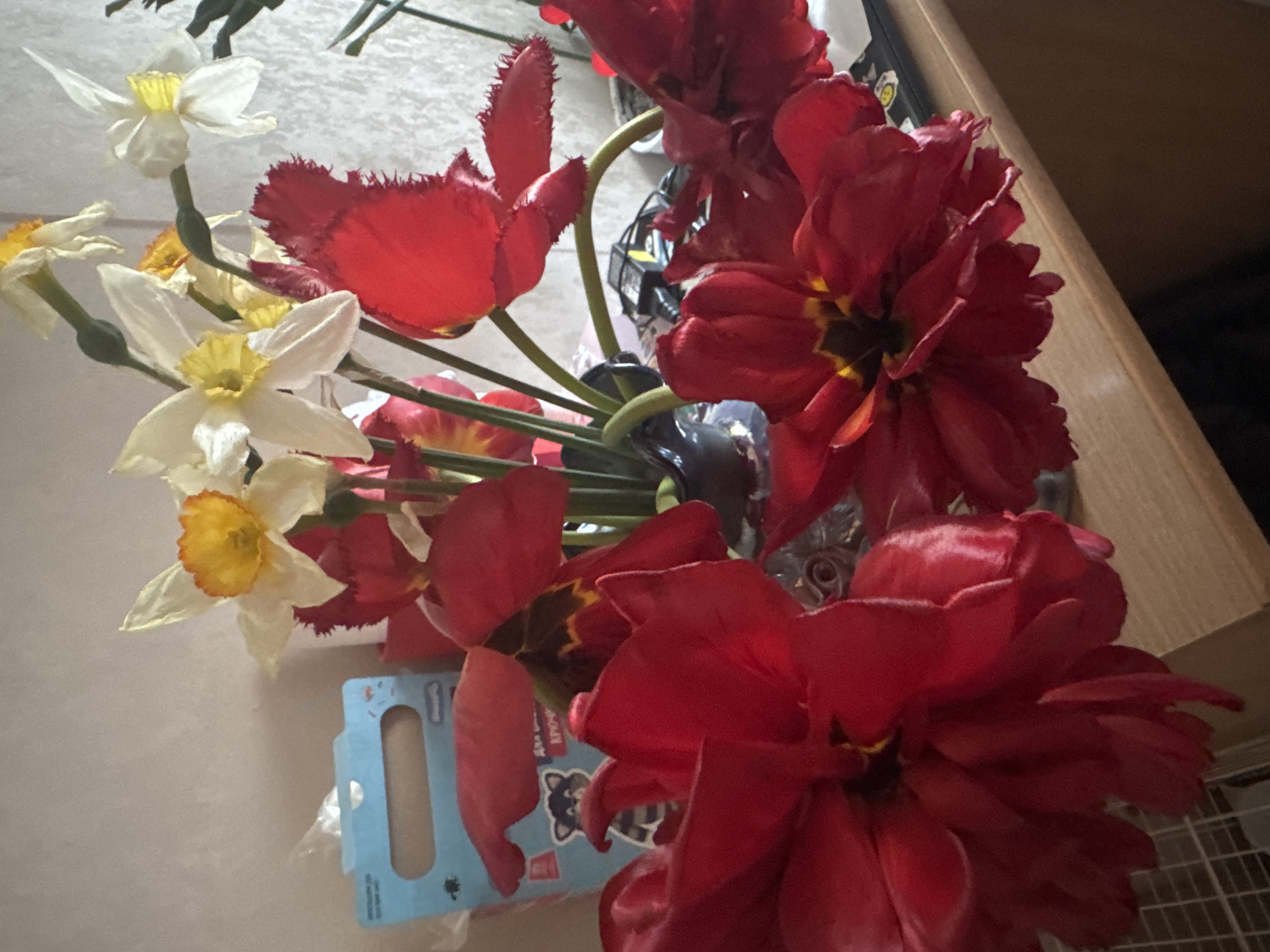 The image shows a floral arrangement in a vase. The bouquet contains red tulips, some with fringed petals, and white and yellow daffodils. The arrangement is placed against a light-colored wall and a wooden surface. A blue box with a cartoon raccoon is visible near the flowers. The location of the image is not discernible.