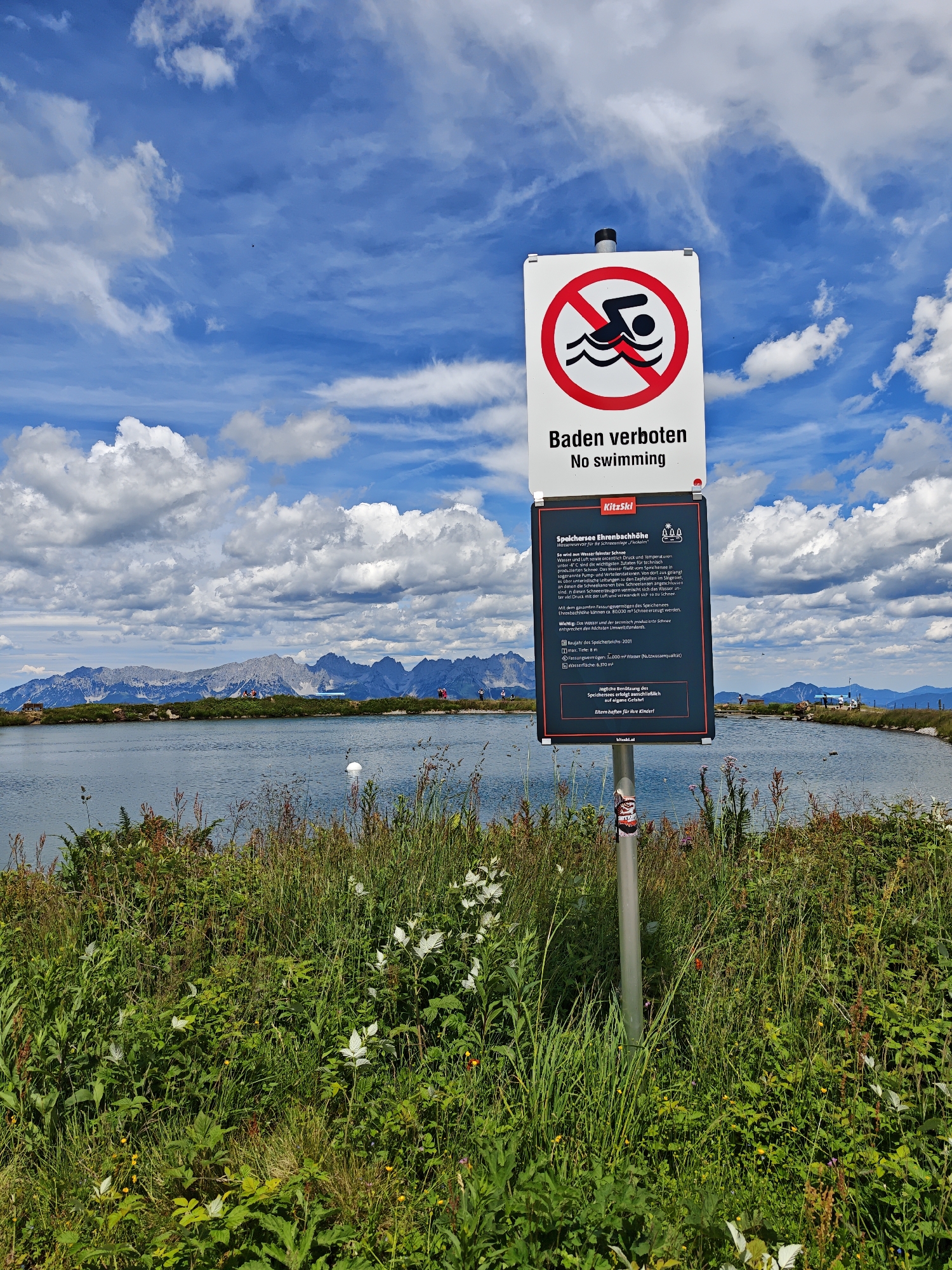 This media file captures a scenic view of a mountain lake in Kirchberg in Tirol, Austria, under a partly cloudy blue sky. In the foreground, a sign with a red circle and a diagonal line over a swimmer, along with German and English text "Baden verboten / No swimming", warns against entering the water. Below this, a detailed informational sign displays "KitzSkl" and provides technical specifications about the "Speichersee Ehrenbachhöhe" (Ehrenbachhöhe Reservoir), including its purpose for snowmaking in the ski resort, its dimensions, and a disclaimer about using the lake at one's own risk. The lake itself is calm and reflects the sky, with lush green grass and wildflowers growing along its banks. In the background, a range of majestic, snow-capped mountains stretches across the horizon, suggesting a high-altitude location. Several people are visible on a grassy ridge on the far side of the lake, possibly enjoying the view or preparing for outdoor activities. The overall impression is one of natural beauty and recreational infrastructure in a mountainous region. The weather appears to be pleasant, with a mix of sun and clouds, typical of a summer day in the Alps.