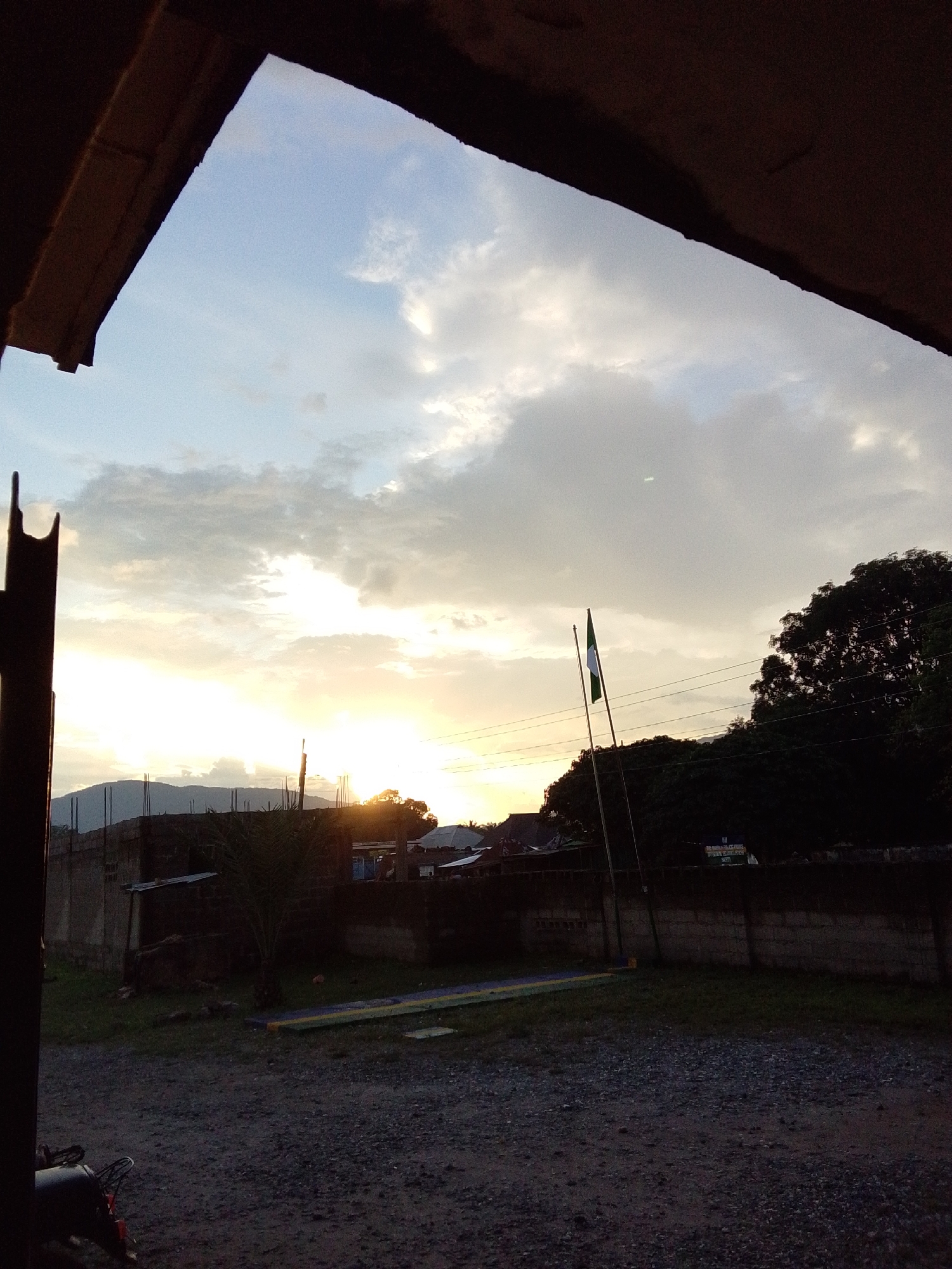 The image depicts a view through an overhang, looking out at a landscape at dusk. The sky is filled with a mixture of light clouds and the warm glow of the setting sun, which is partially obscured by a mountain range in the distance. In the middle ground, there is a low-rise building with a concrete wall and a partially constructed structure behind it. Two flagpoles stand in front of a cluster of trees, one flying a flag that appears to be green and white. The foreground is a gravelly area with some patches of grass. To the left, the handlebars and front wheel of a motorcycle are visible, suggesting a private or residential area. The scene appears to be a quiet, perhaps rural or semi-urban setting within Serti, Nigeria, characterized by its natural light conditions and modest infrastructure.